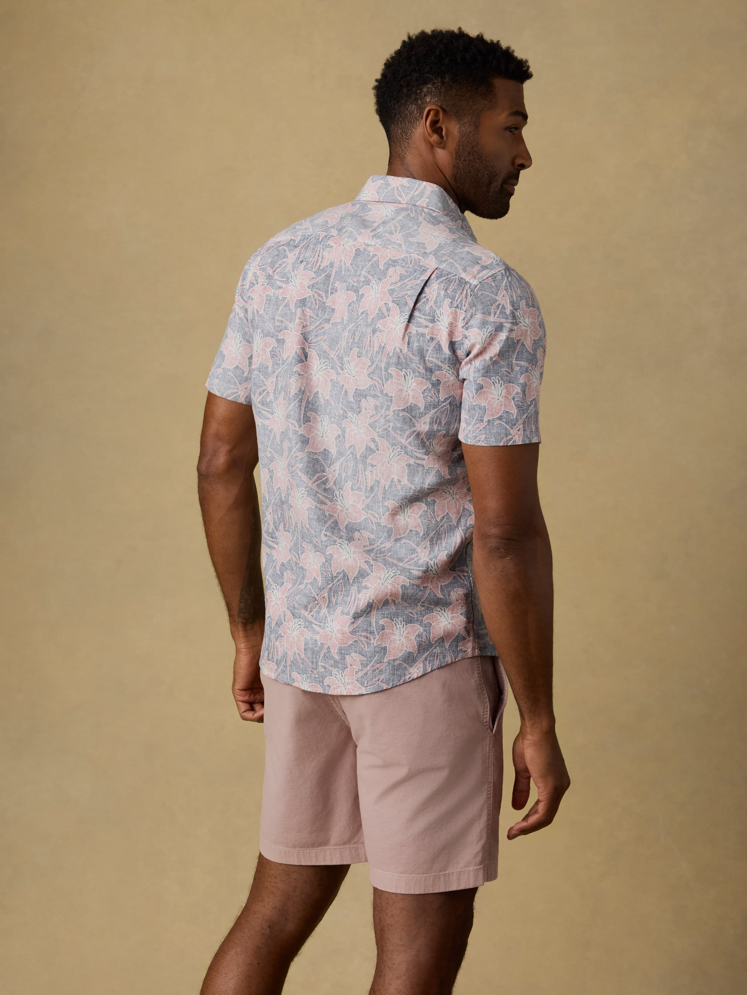 Back view of a man wearing a floral printed short sleeve button down shirt and pink shorts standing against a beige background