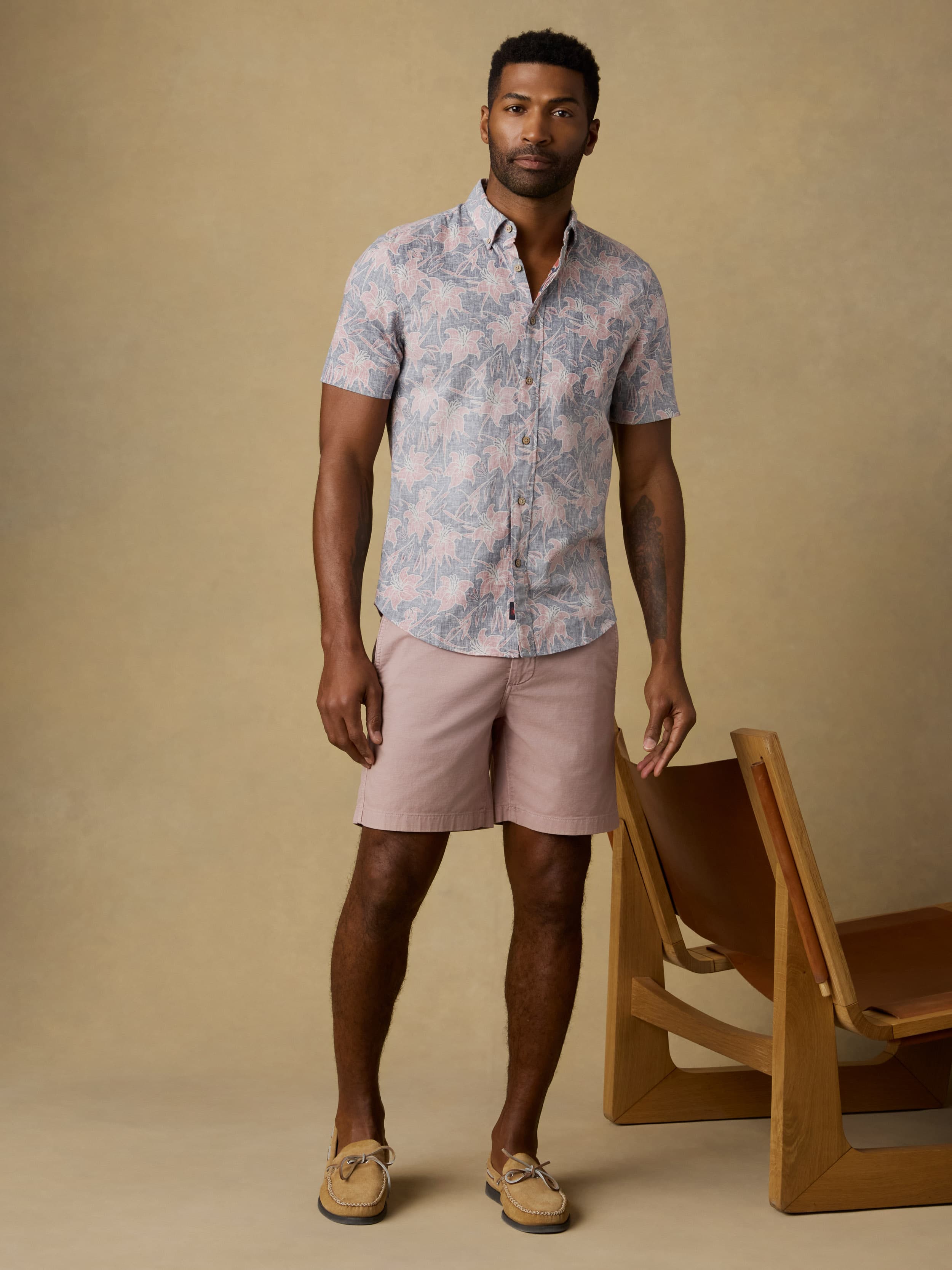 Man wearing a floral printed short sleeve button down shirt and pink shorts standing against a beige background