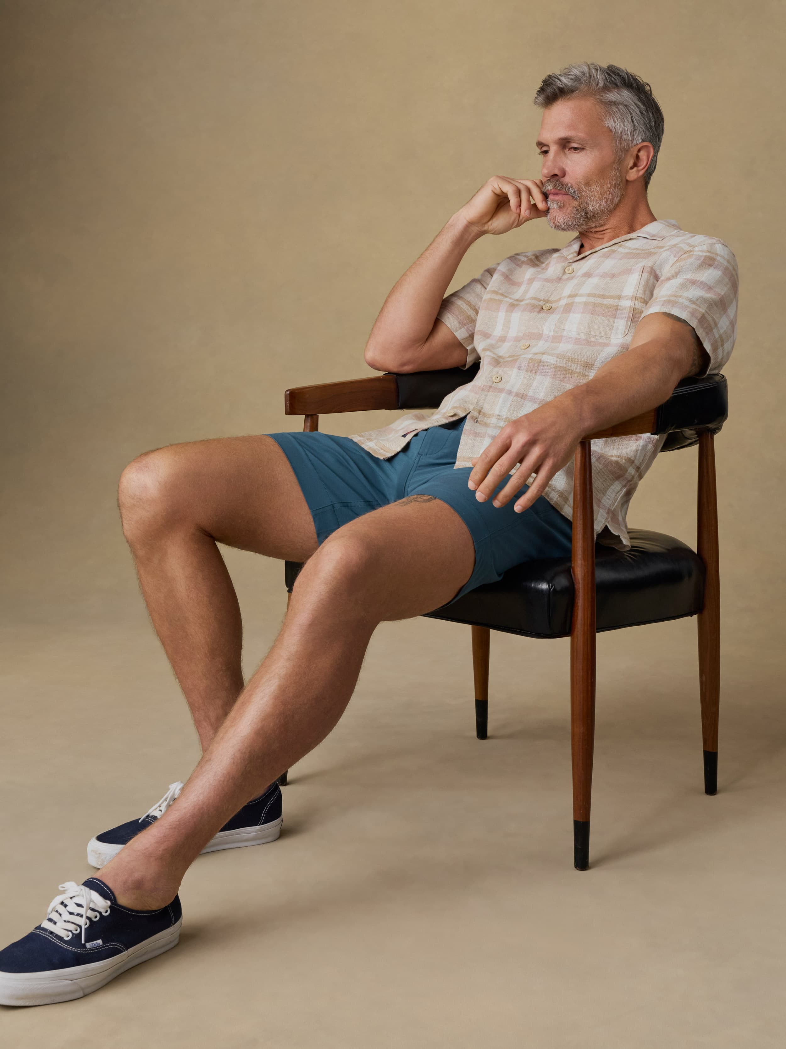 Man sitting on a chair wearing a plaid shirt and blue shorts against a beige background