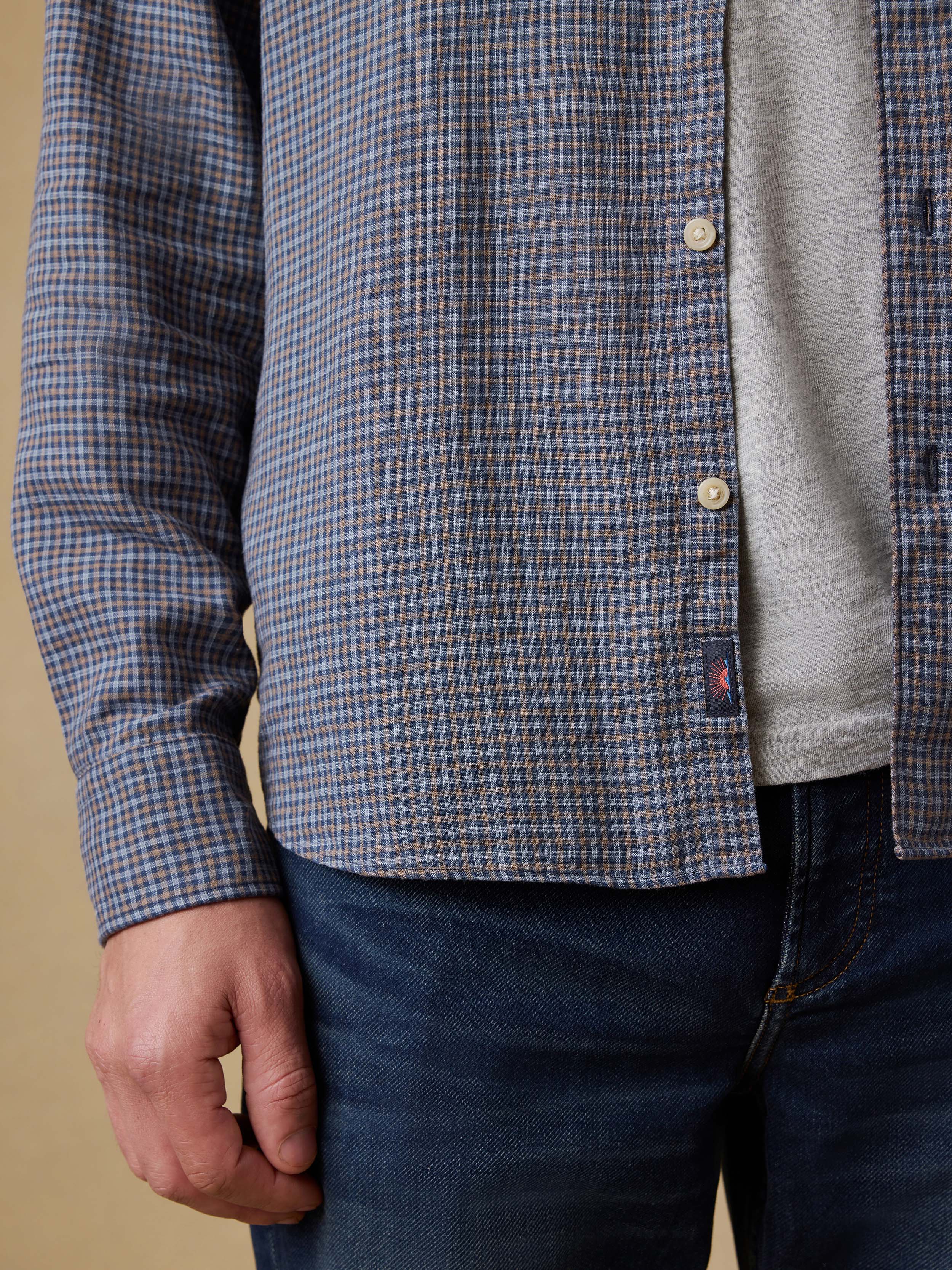 Close-up of a man wearing a blue plaid shirt with a visible Faherty brand logo at the hem over a gray t-shirt with a beige background