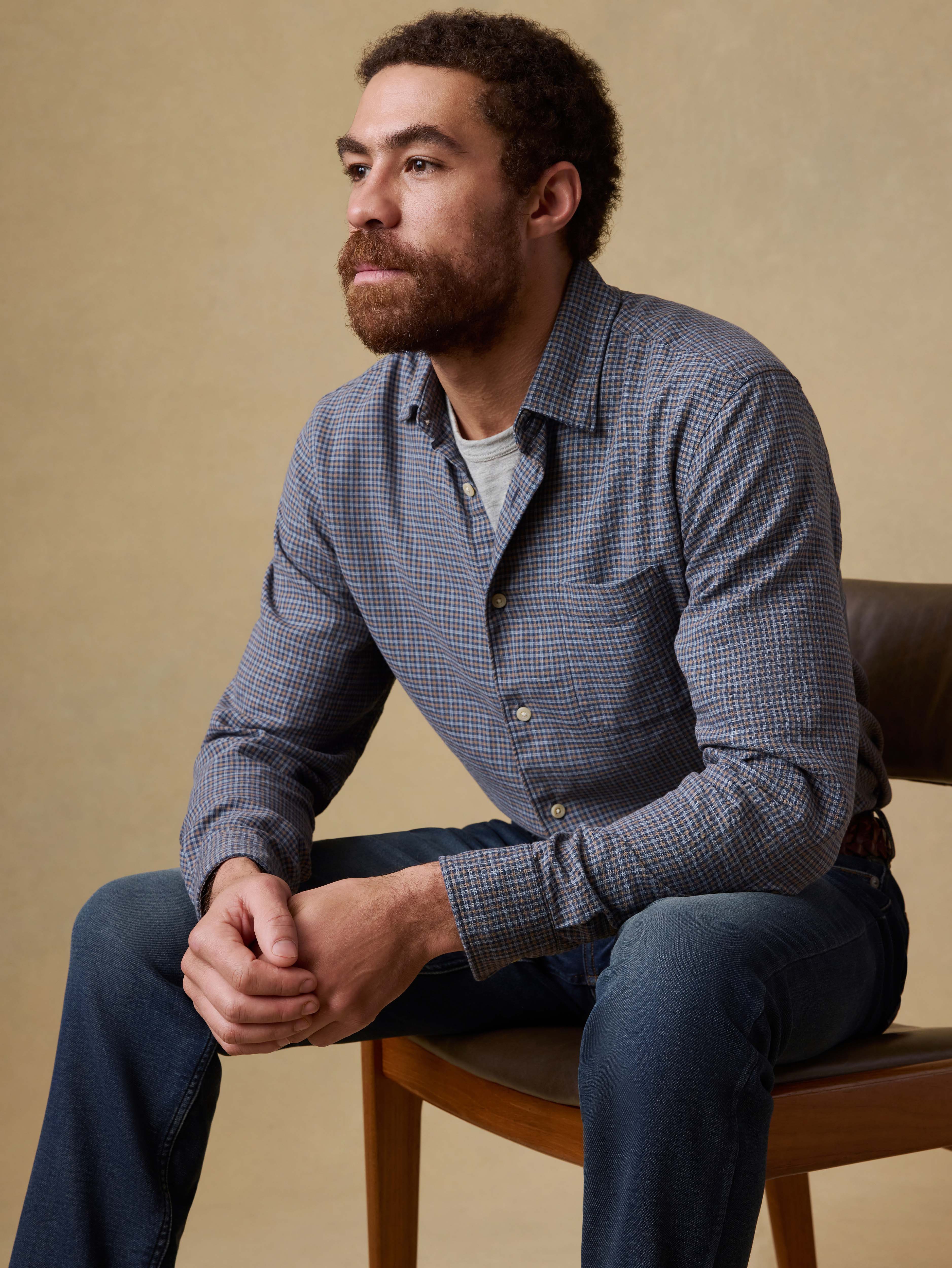 Man sitting on a chair wearing a blue checkered shirt over a gray t-shirt and blue jeans against a beige background