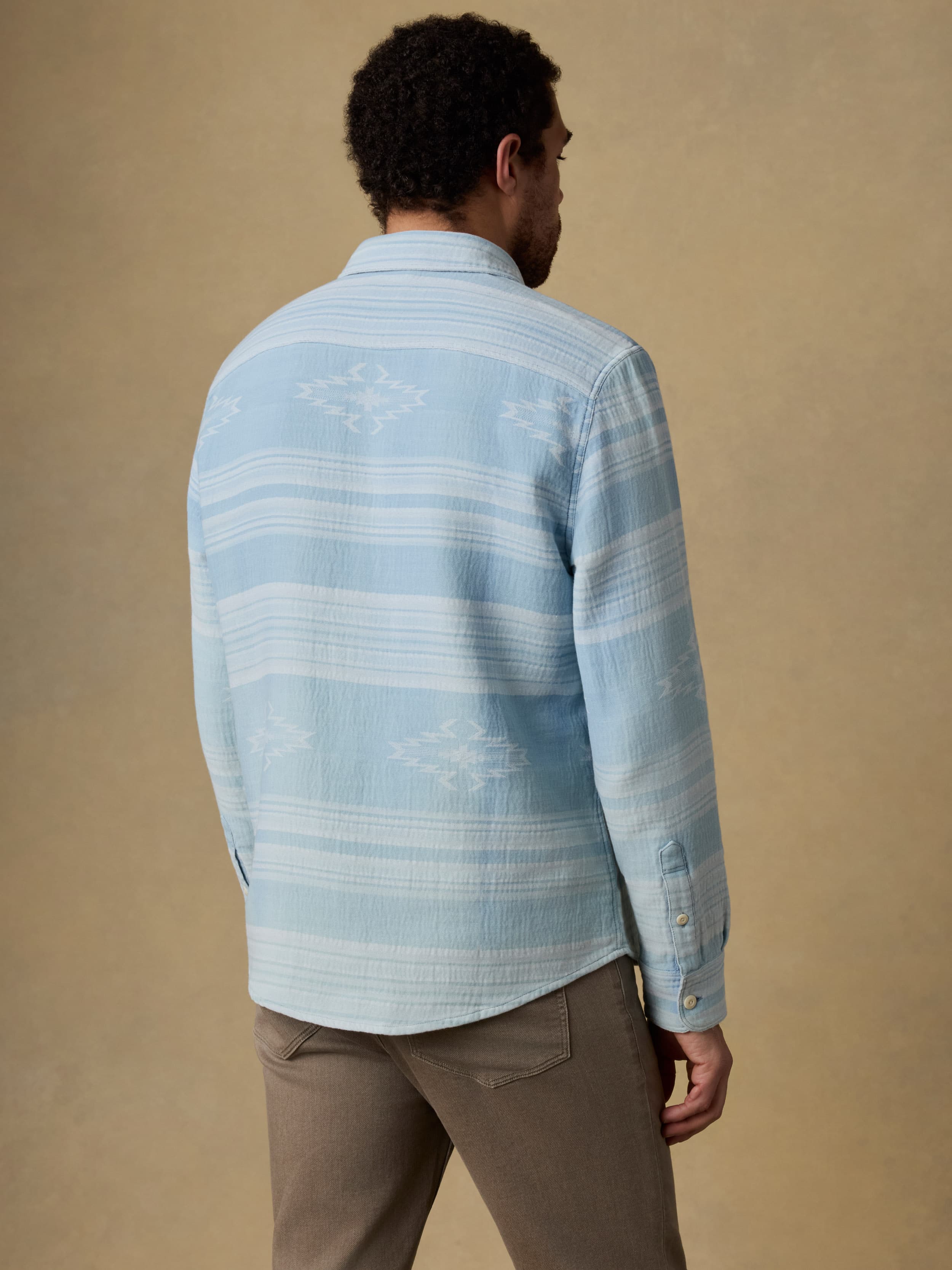 Back view of a man wearing a light indigo button down shirt and brown jeans against a beige background