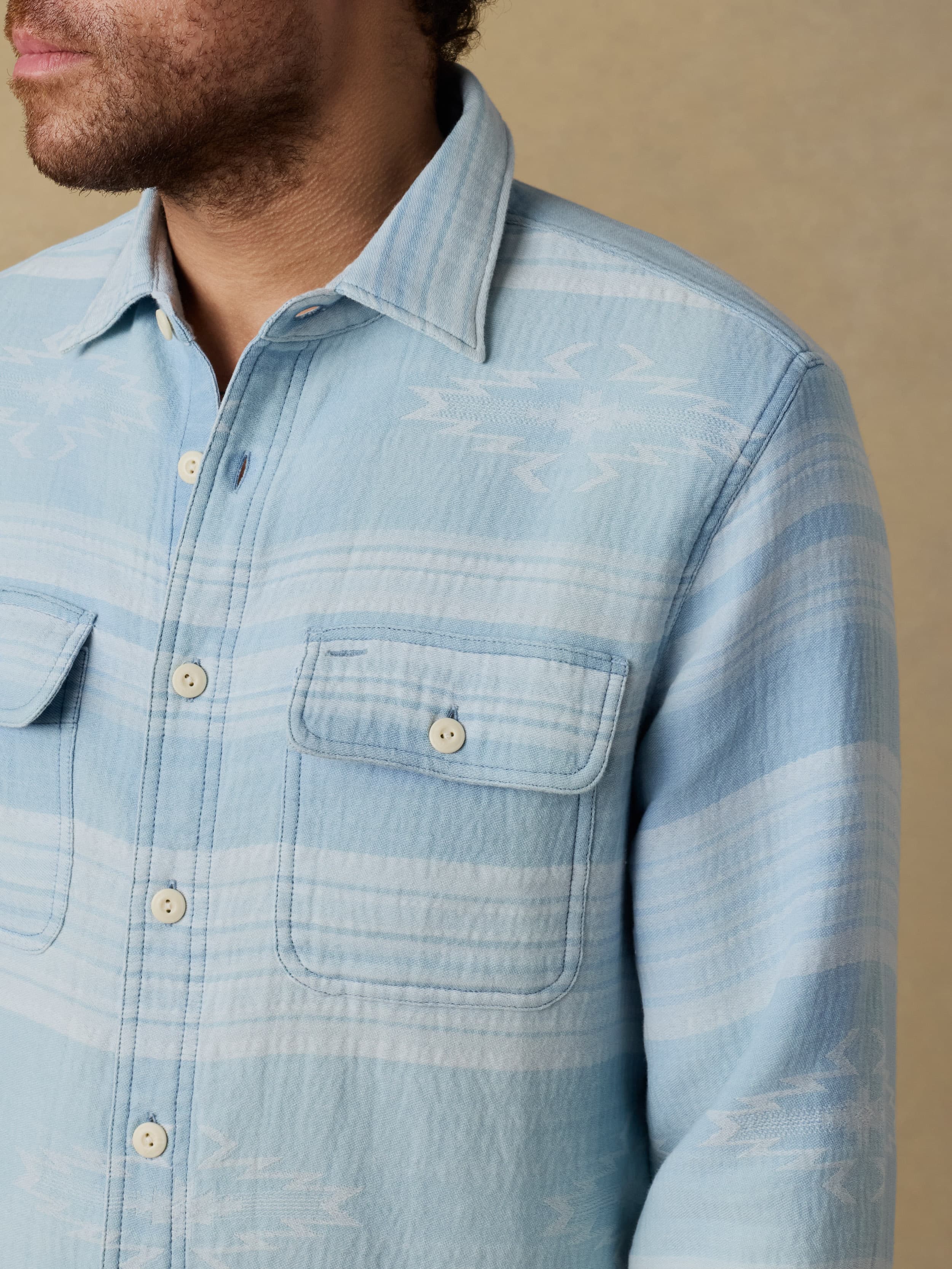 Close up shot of a man wearing a light indigo button down shirt against a beige background