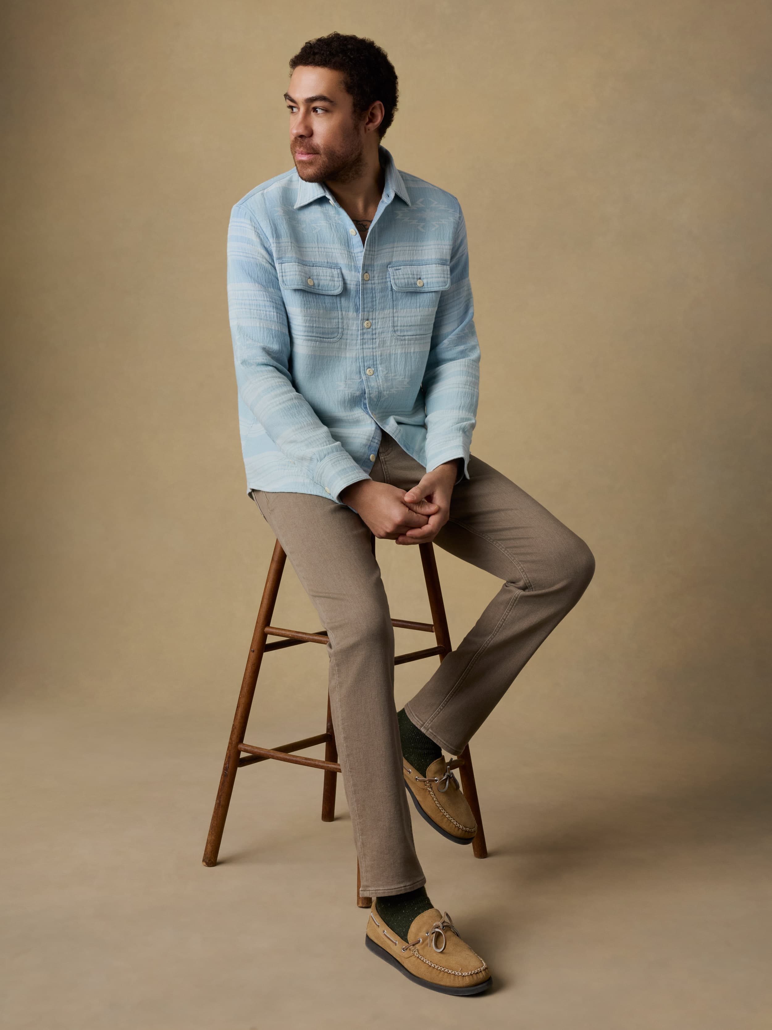 Man sitting on a wooden stool wearing a light indigo button down shirt and brown jeans against a beige background