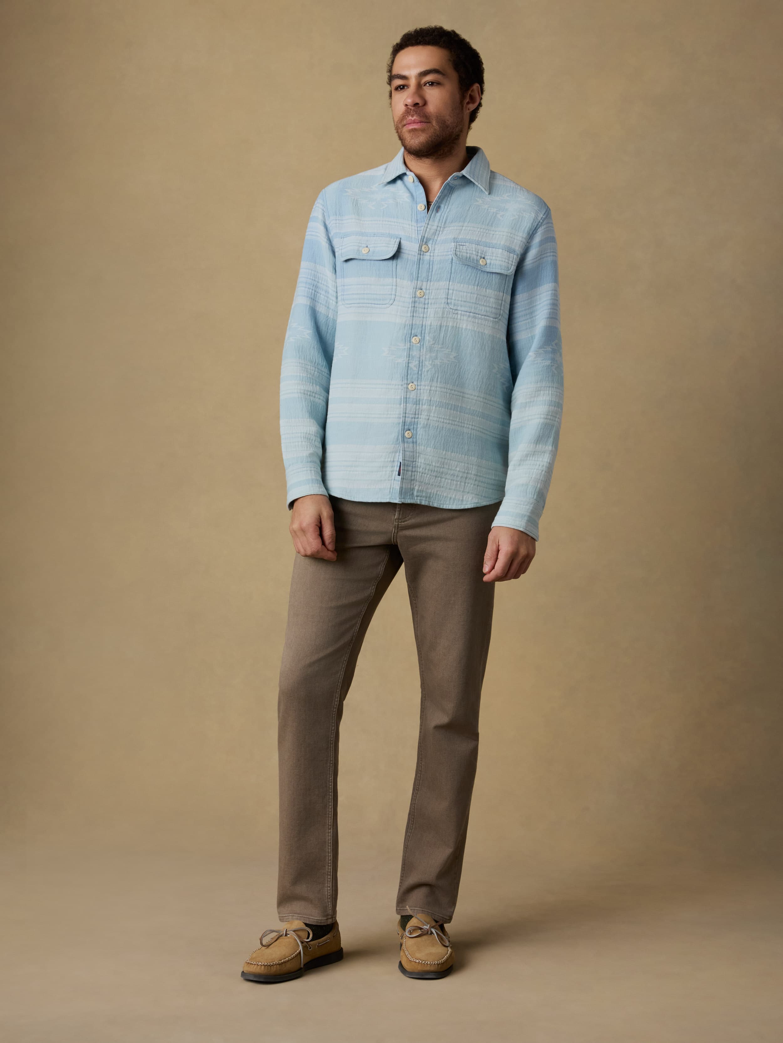 Man wearing a light indigo button down shirt and brown jeans against a beige background