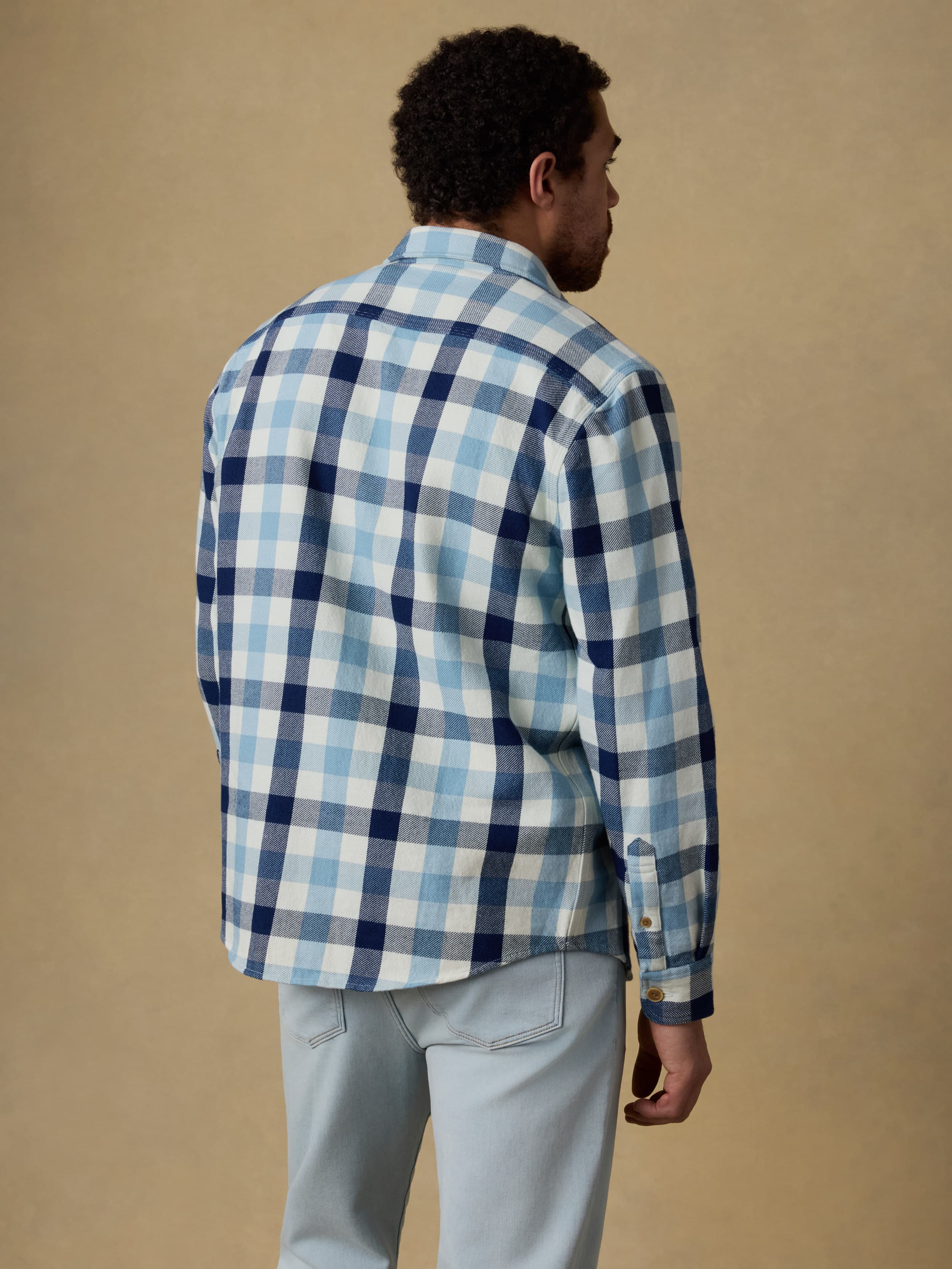 Back view of a man wearing a blue and white plaid long sleeve button up shirt over a white t-shirt and blue jeans on a beige background