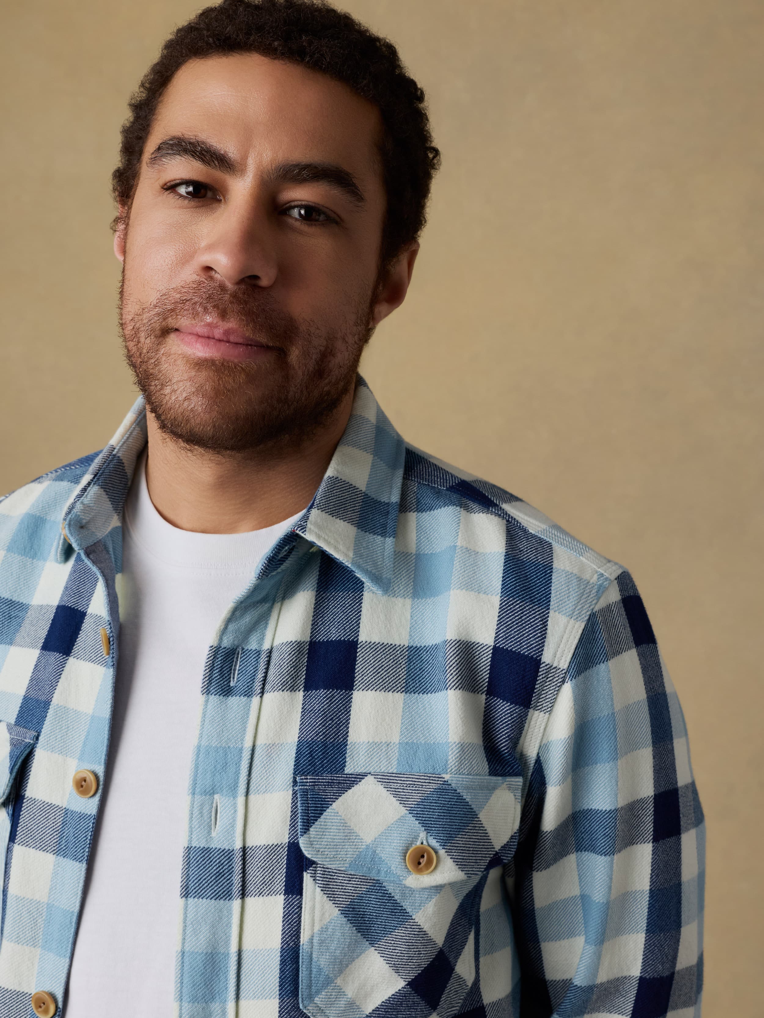 Detail shot of a man wearing a blue and white plaid long sleeve button up shirt over a white t-shirt on a beige background