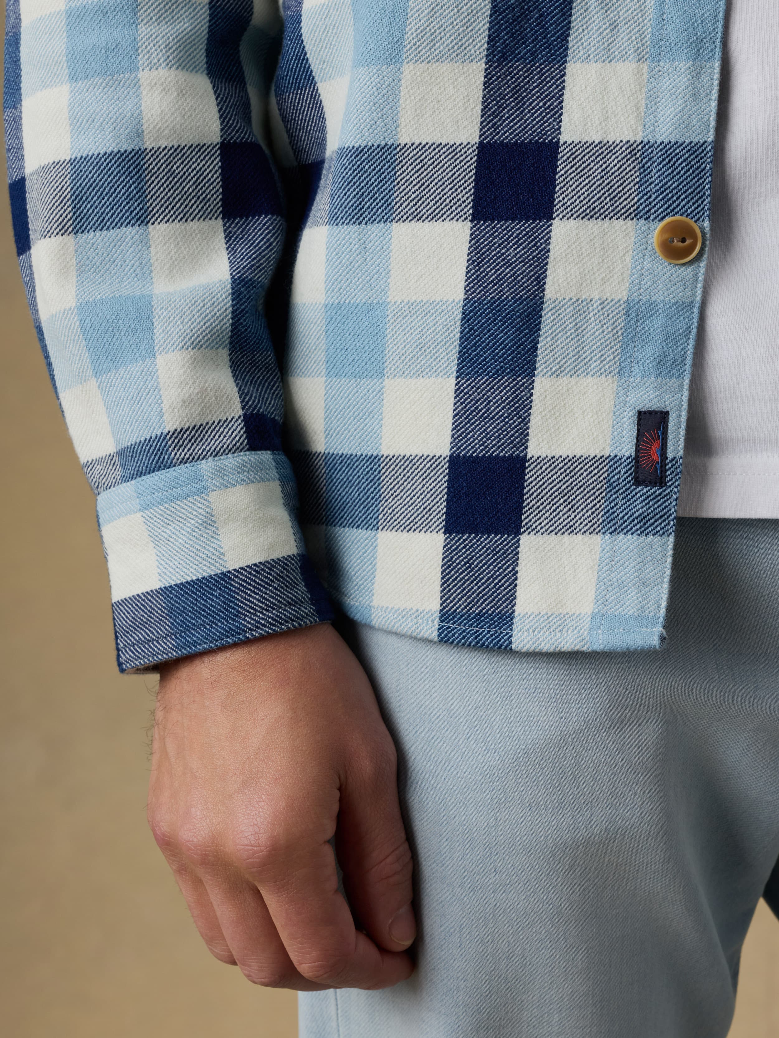 Close up shot of a man wearing a blue and white plaid long sleeve button up shirt with a visible Faherty brand logo near the hem and blue jeans on a beige background
