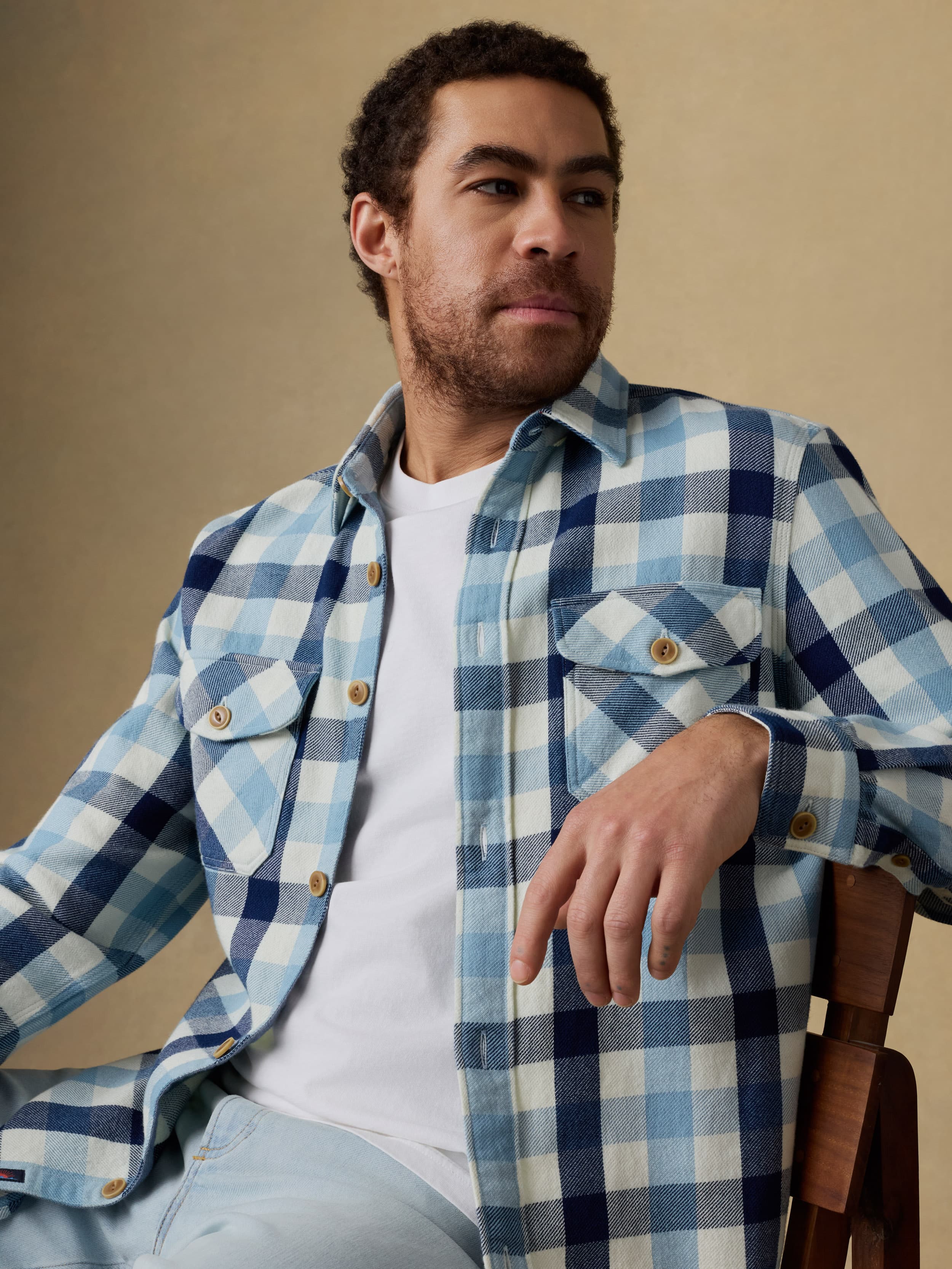 Man sitting on a wooden chair wearing a blue and white plaid long sleeve button up shirt over a white t-shirt and blue jeans on a beige background
