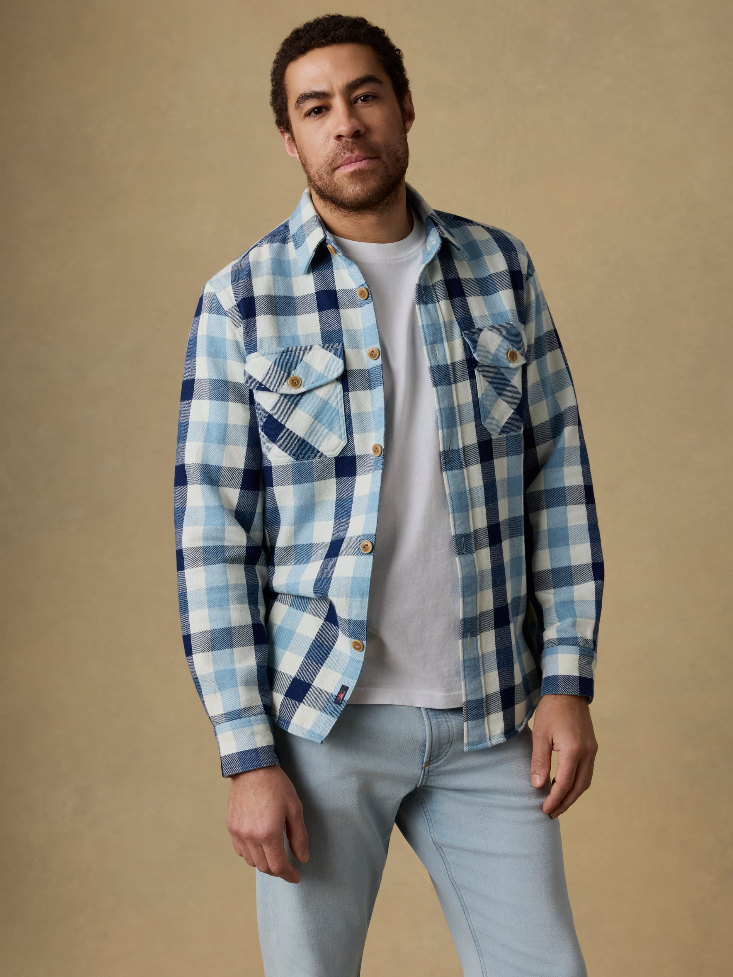Man wearing a blue and white plaid long sleeve button up shirt over a white t-shirt and blue jeans on a beige background
