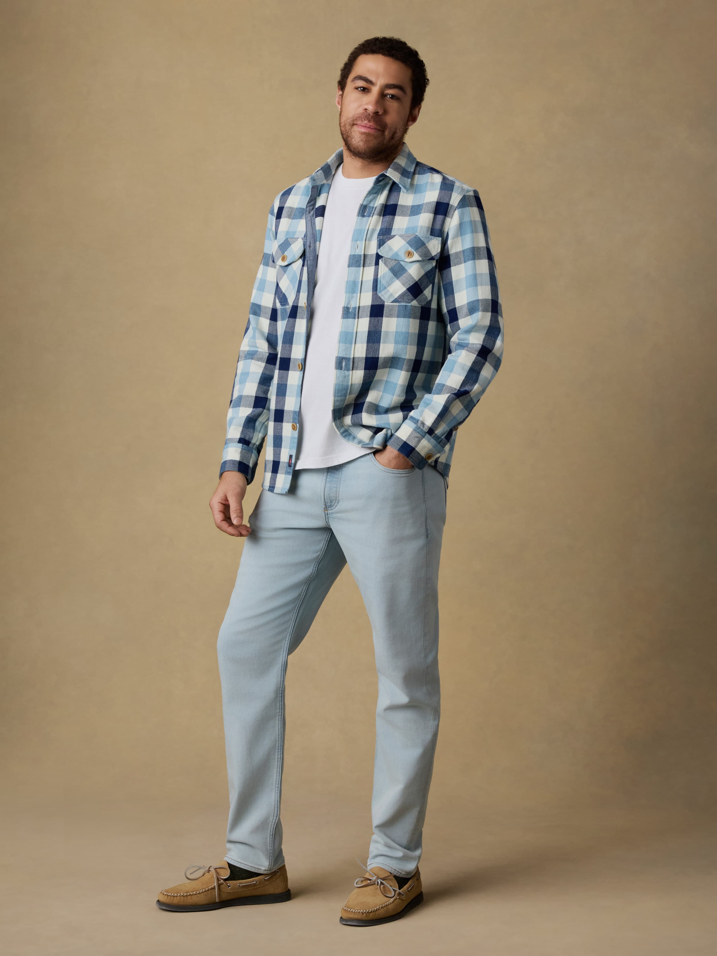 Man wearing a blue and white plaid long sleeve button up shirt over a white t-shirt and blue jeans on a beige background
