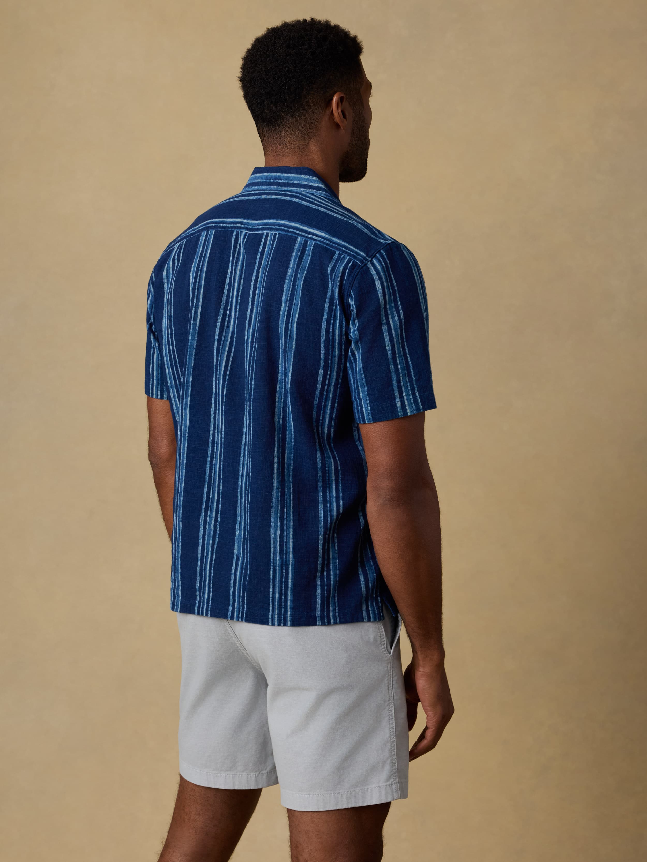 Back view of a man wearing a blue striped short sleeve button down shirt and light grey shorts against a beige background