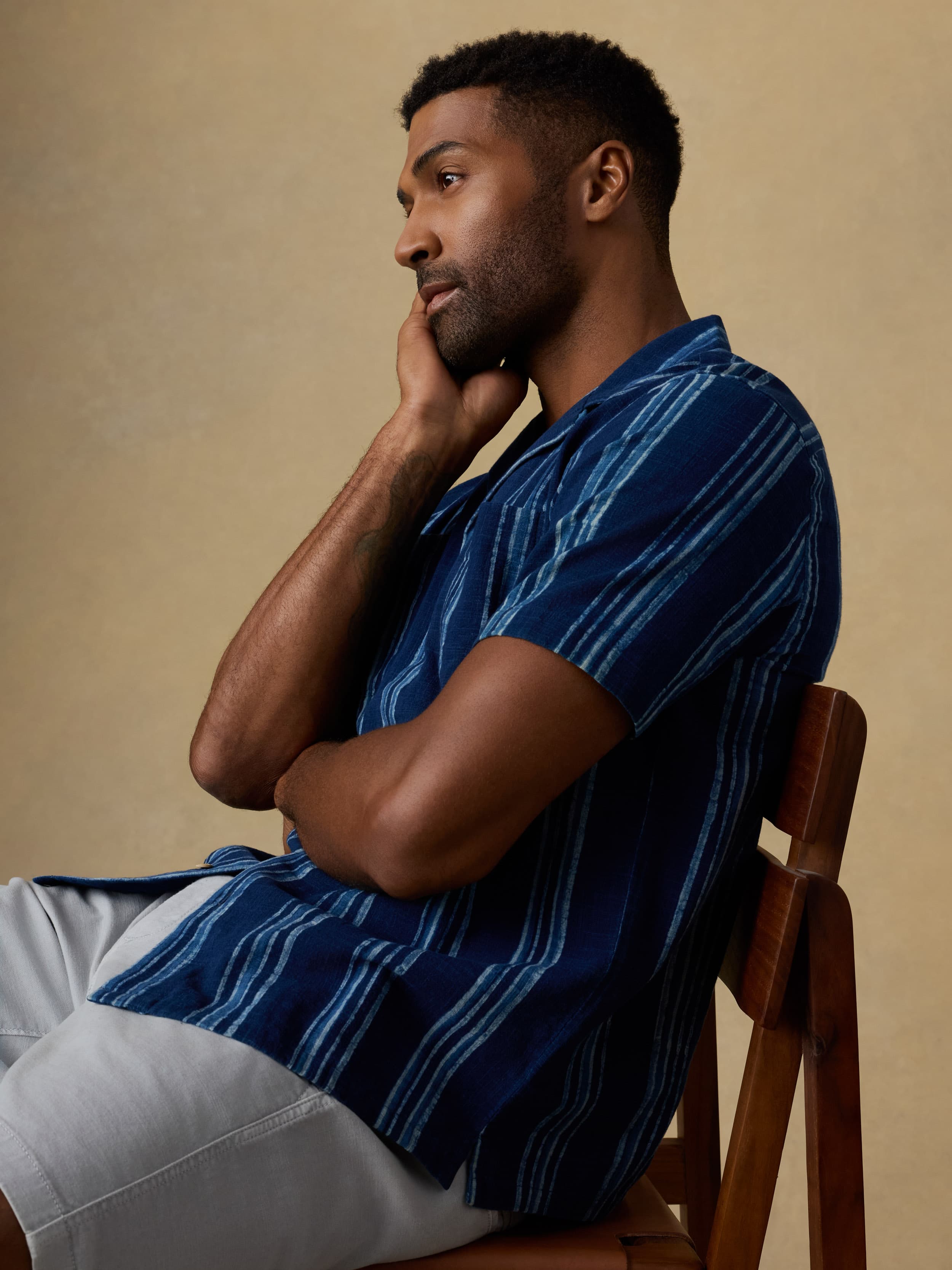 Man sitting on a wooden chair wearing a blue striped short sleeve button down shirt and light grey shorts against a beige background