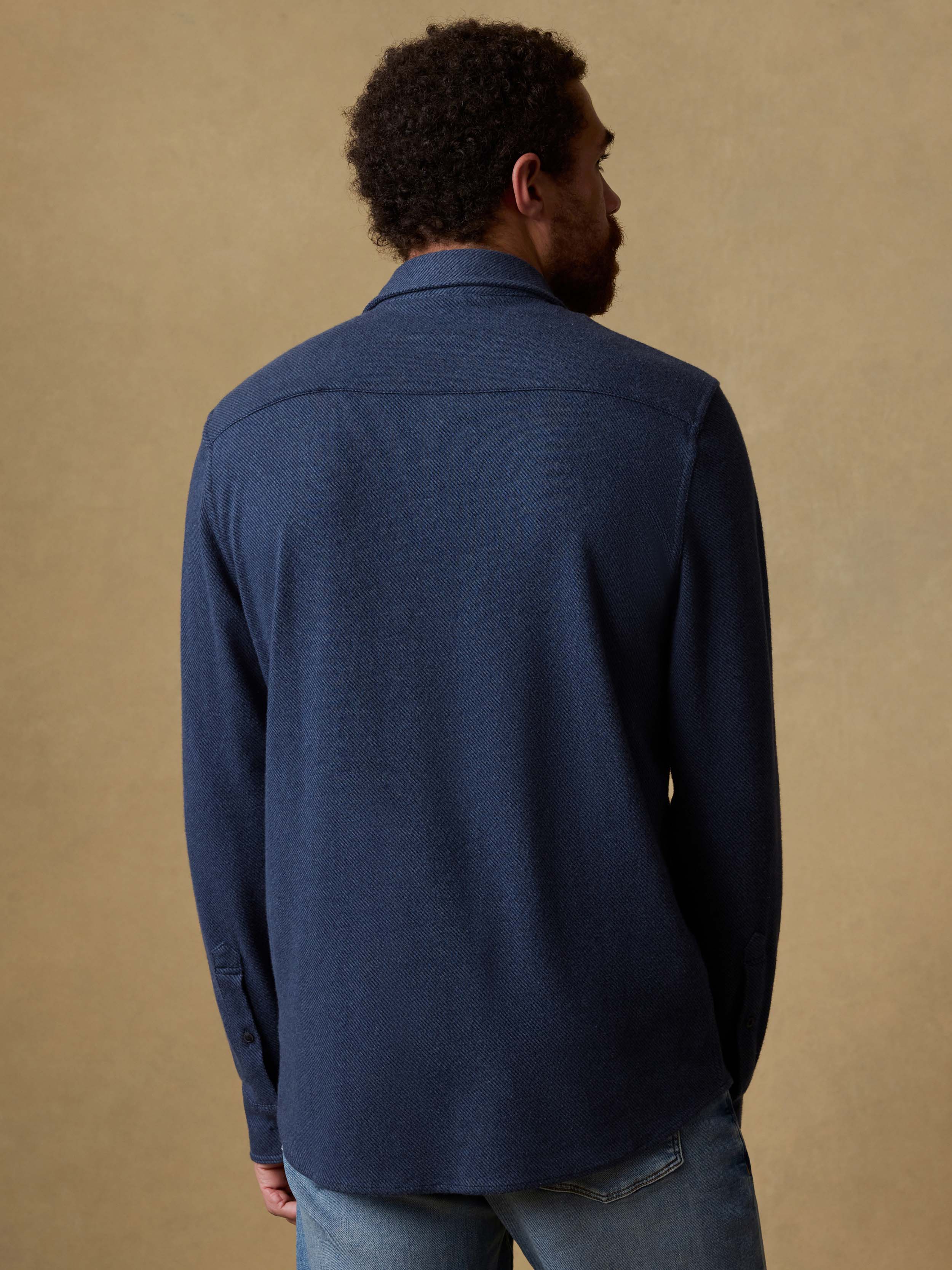 Back view of a man wearing a navy blue button down shirt and jeans against a beige background