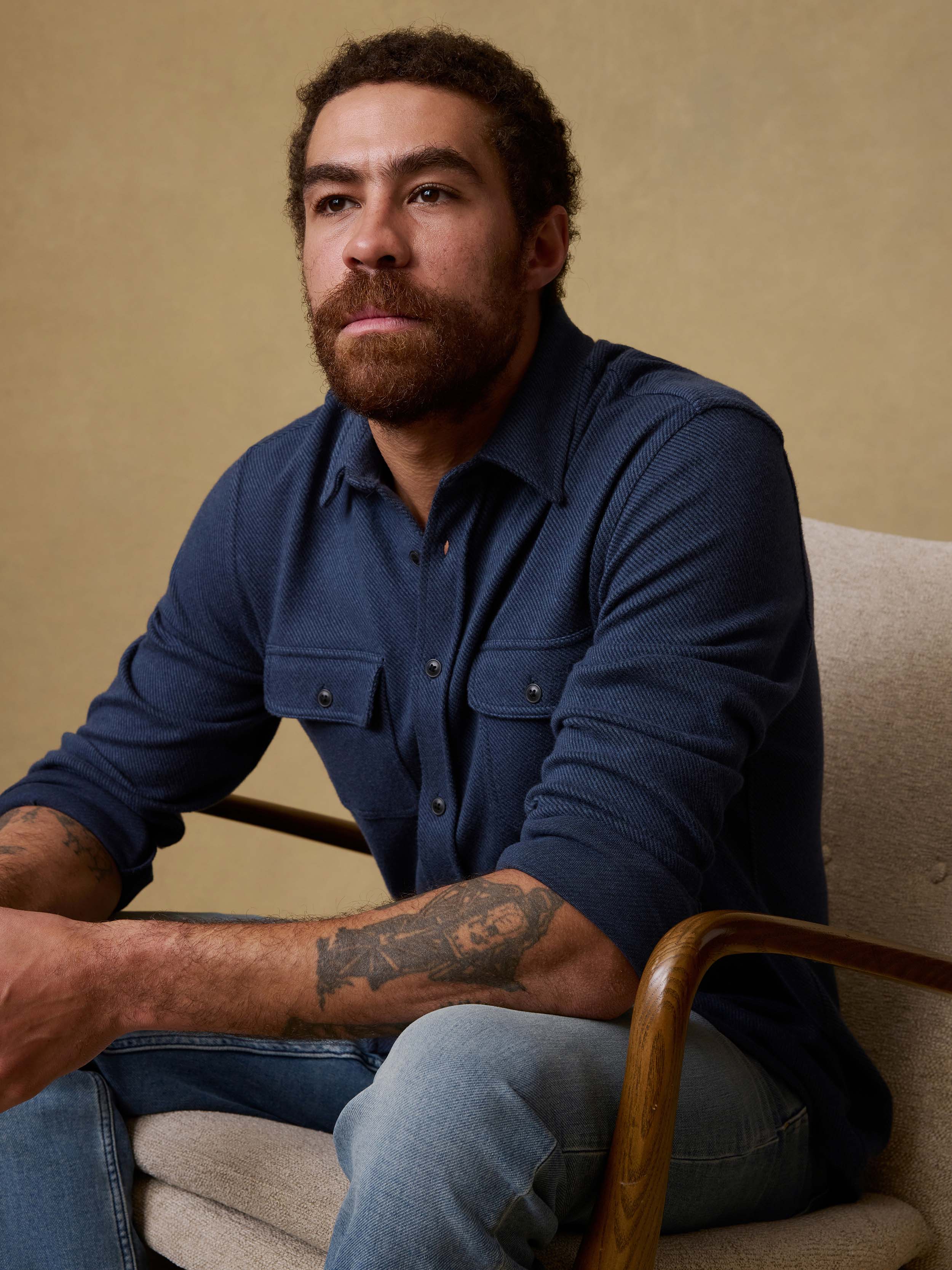Man sitting on a chair wearing a navy blue button down shirt and jeans against a beige background