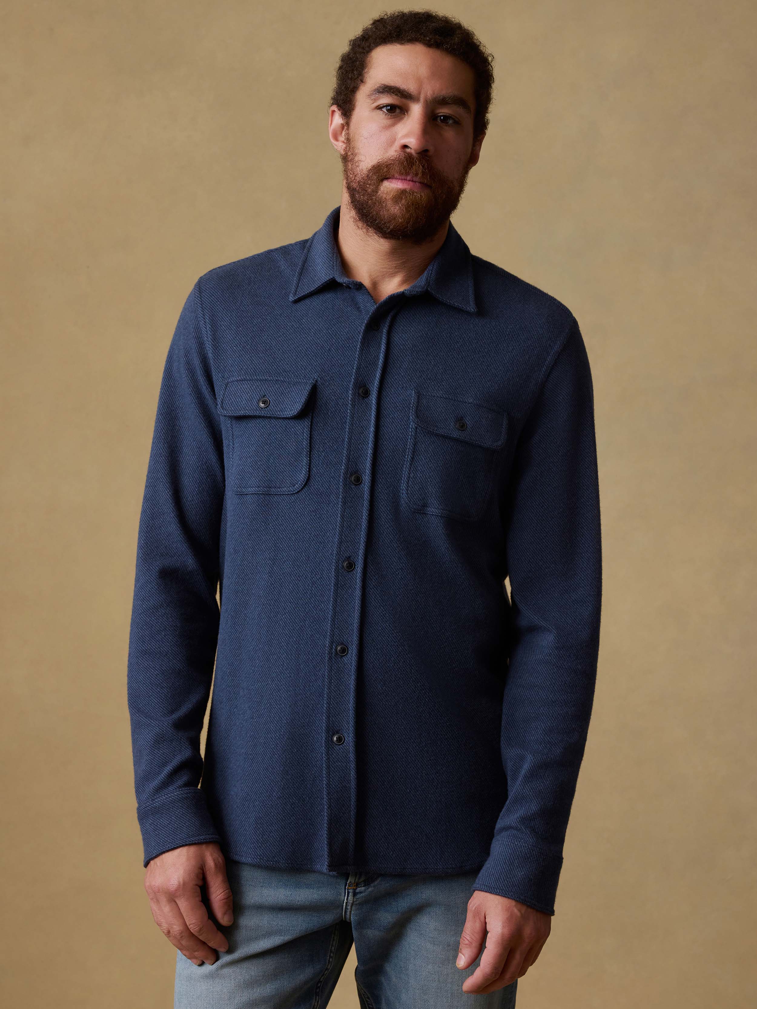 Man wearing a navy blue button down shirt and jeans against a beige background