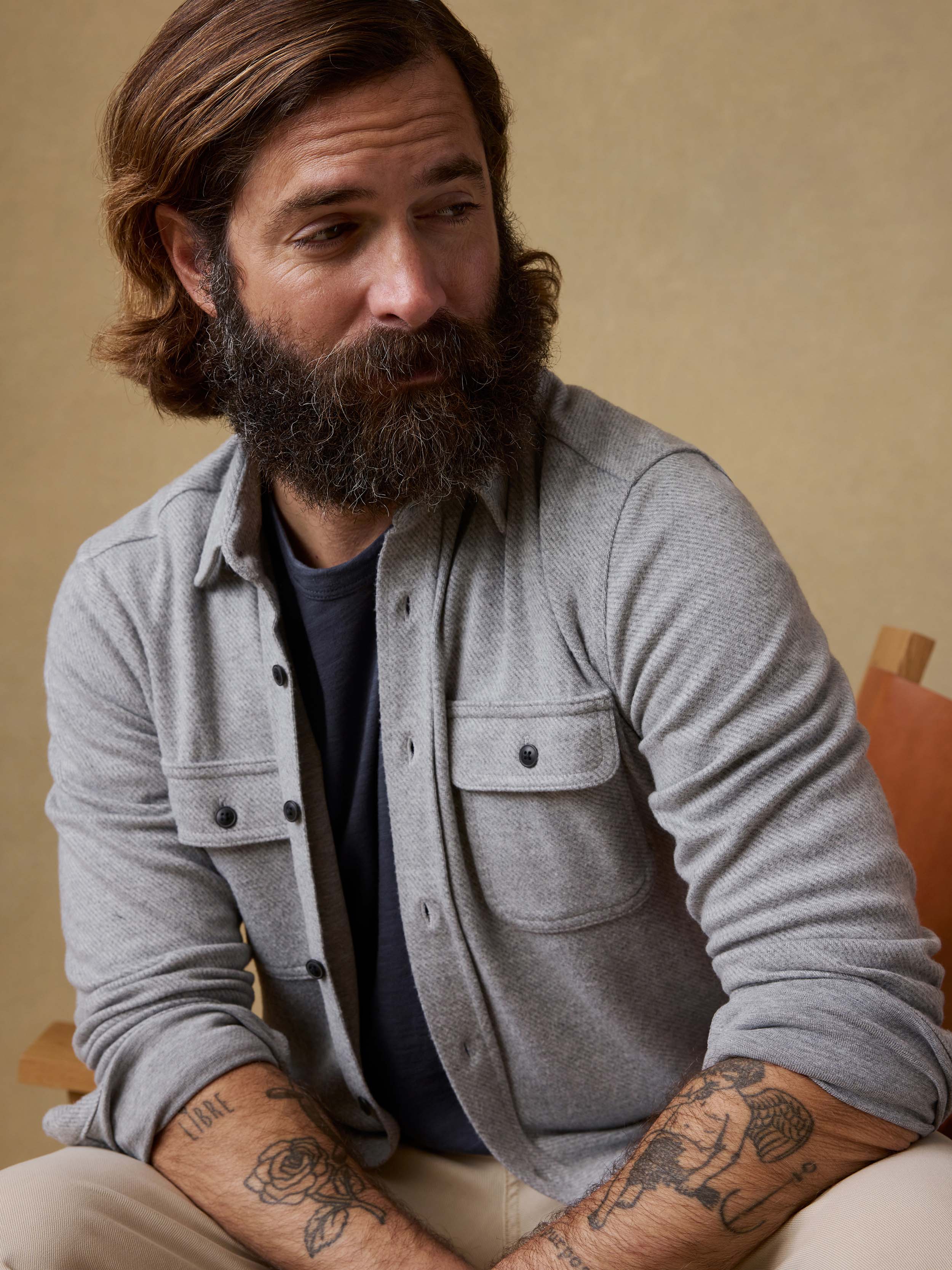 Man sitting on a chair wearing a gray long-sleeve shirt and beige pants against a beige background