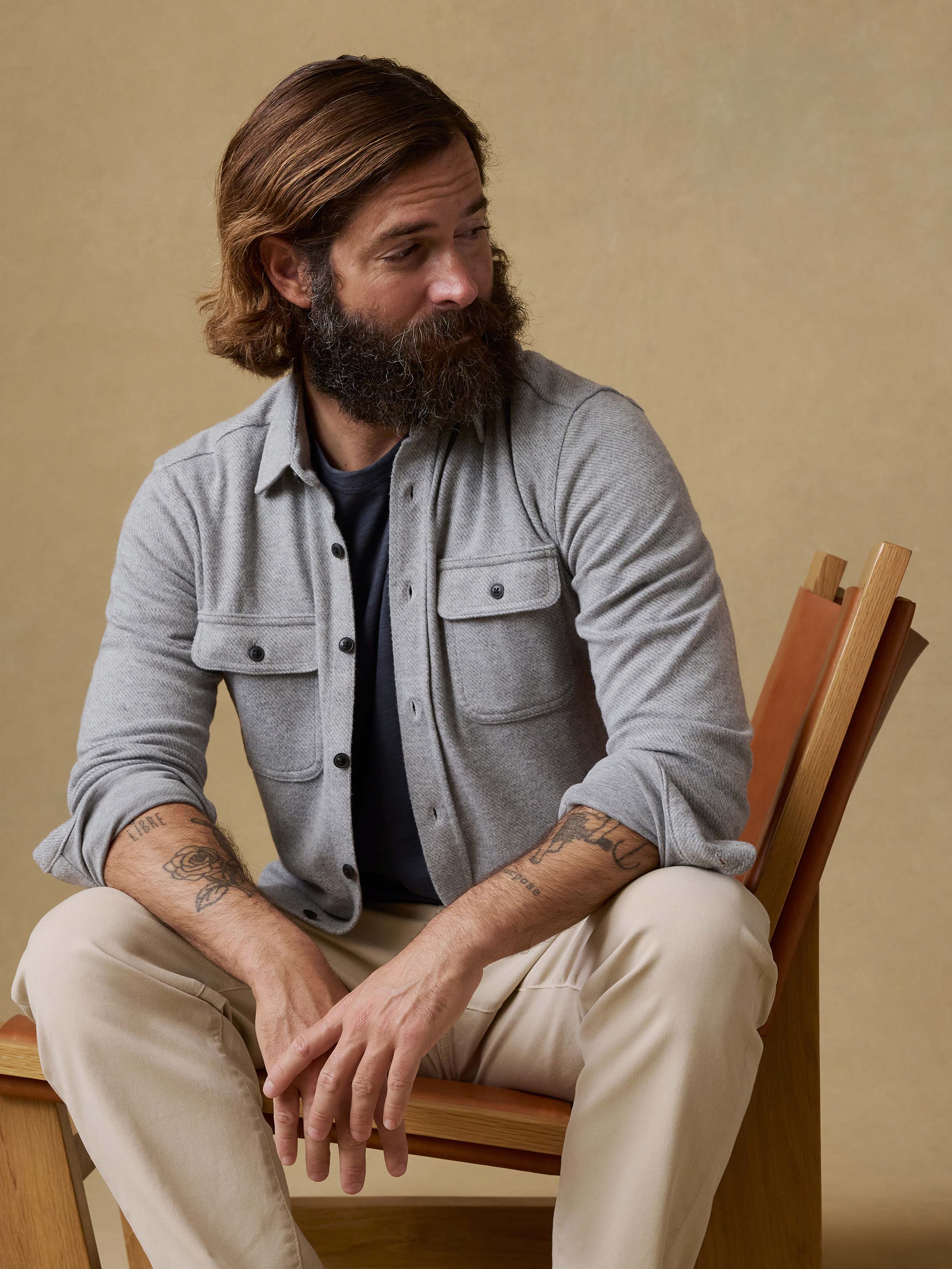 Man sitting on a chair wearing a gray long-sleeve shirt and beige pants against a beige background