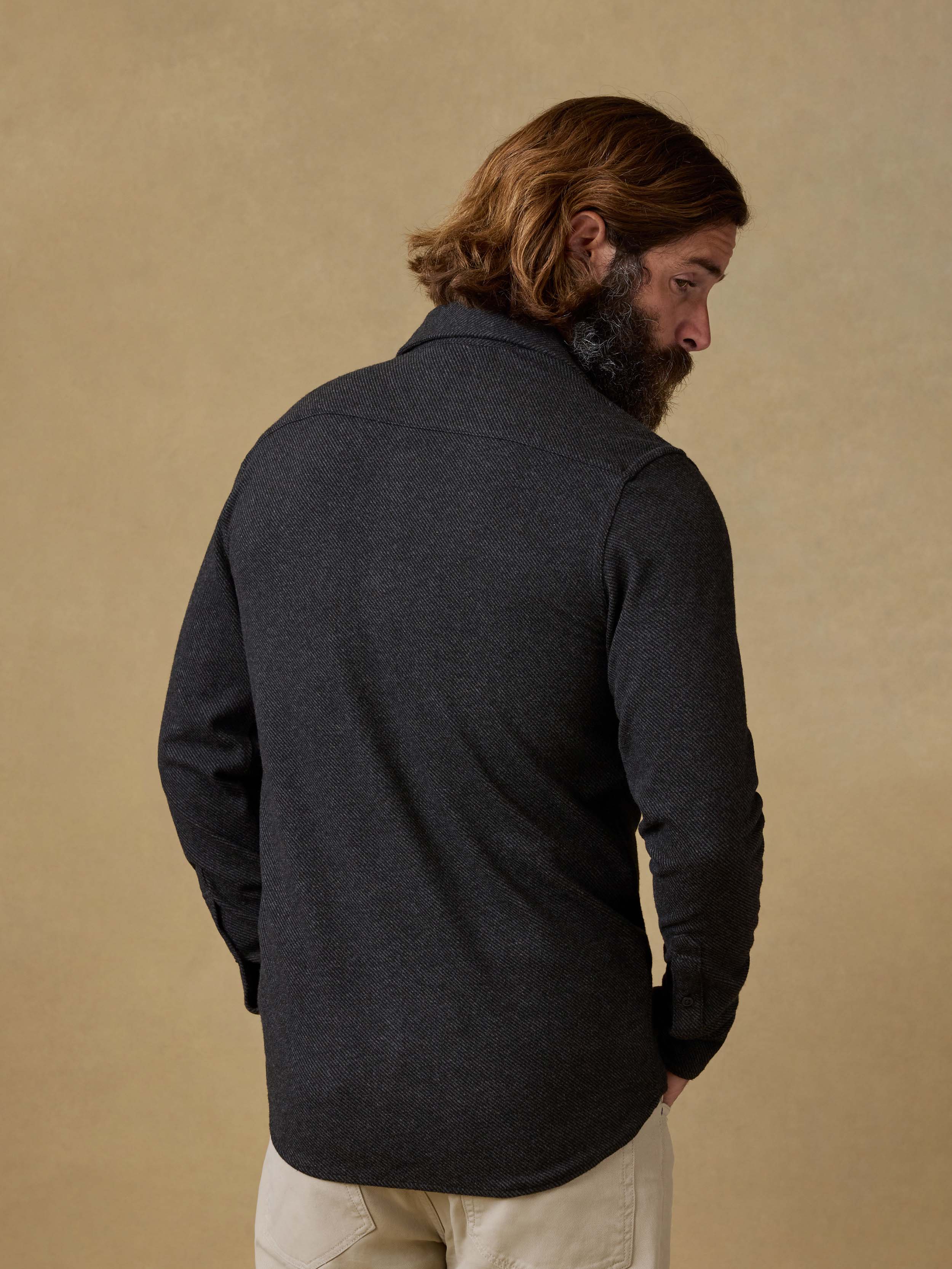 Back view of a man wearing a black button down shirt with beige pants against a beige background