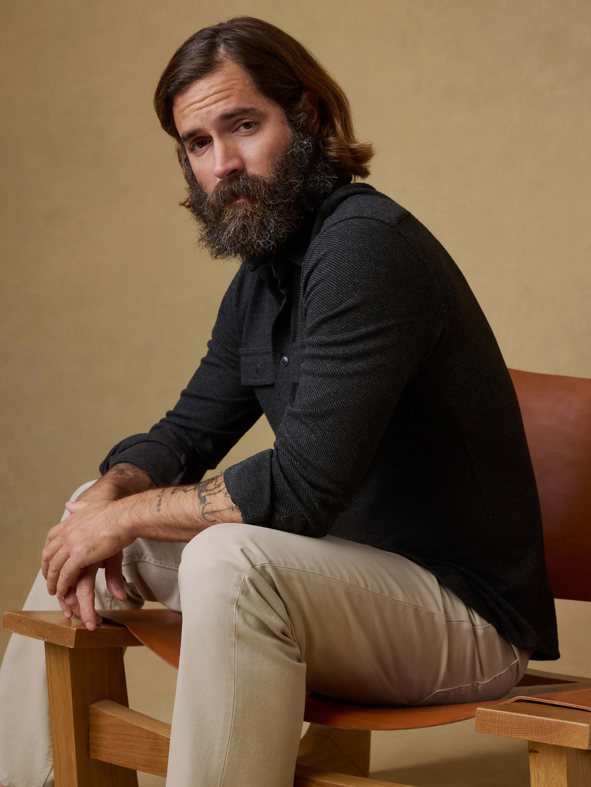 Man sitting on a chair wearing a black button down shirt with beige pants against a beige background