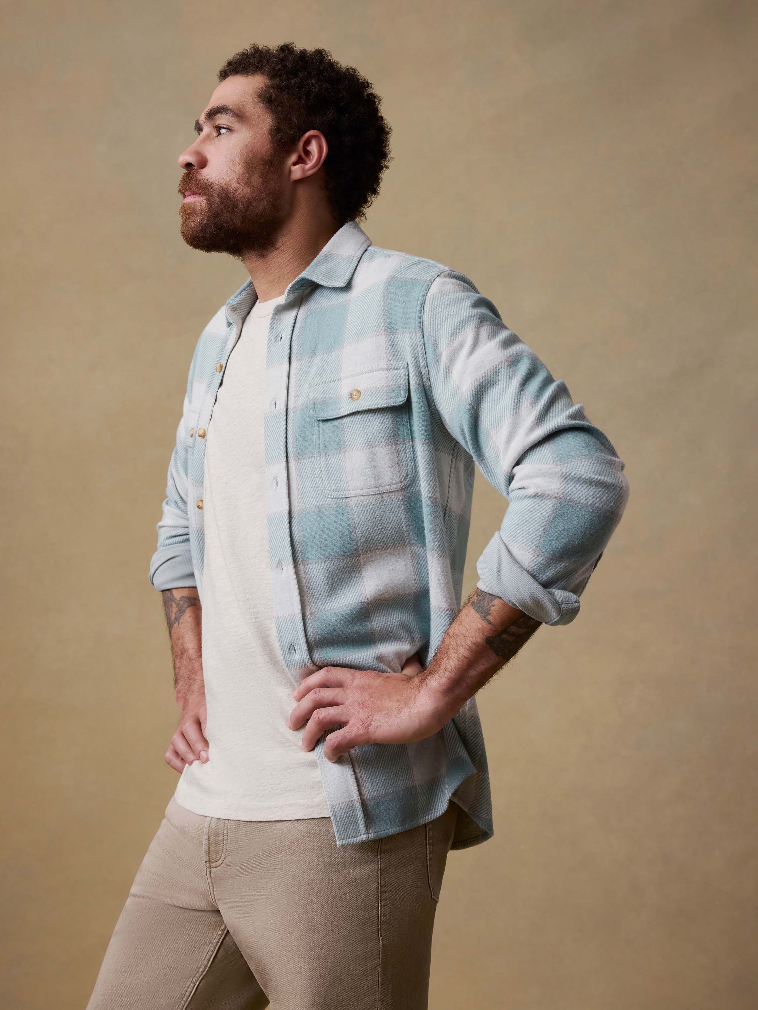 Man wearing a light blue flannel shirt over a white t-shirt with beige pants against a beige background