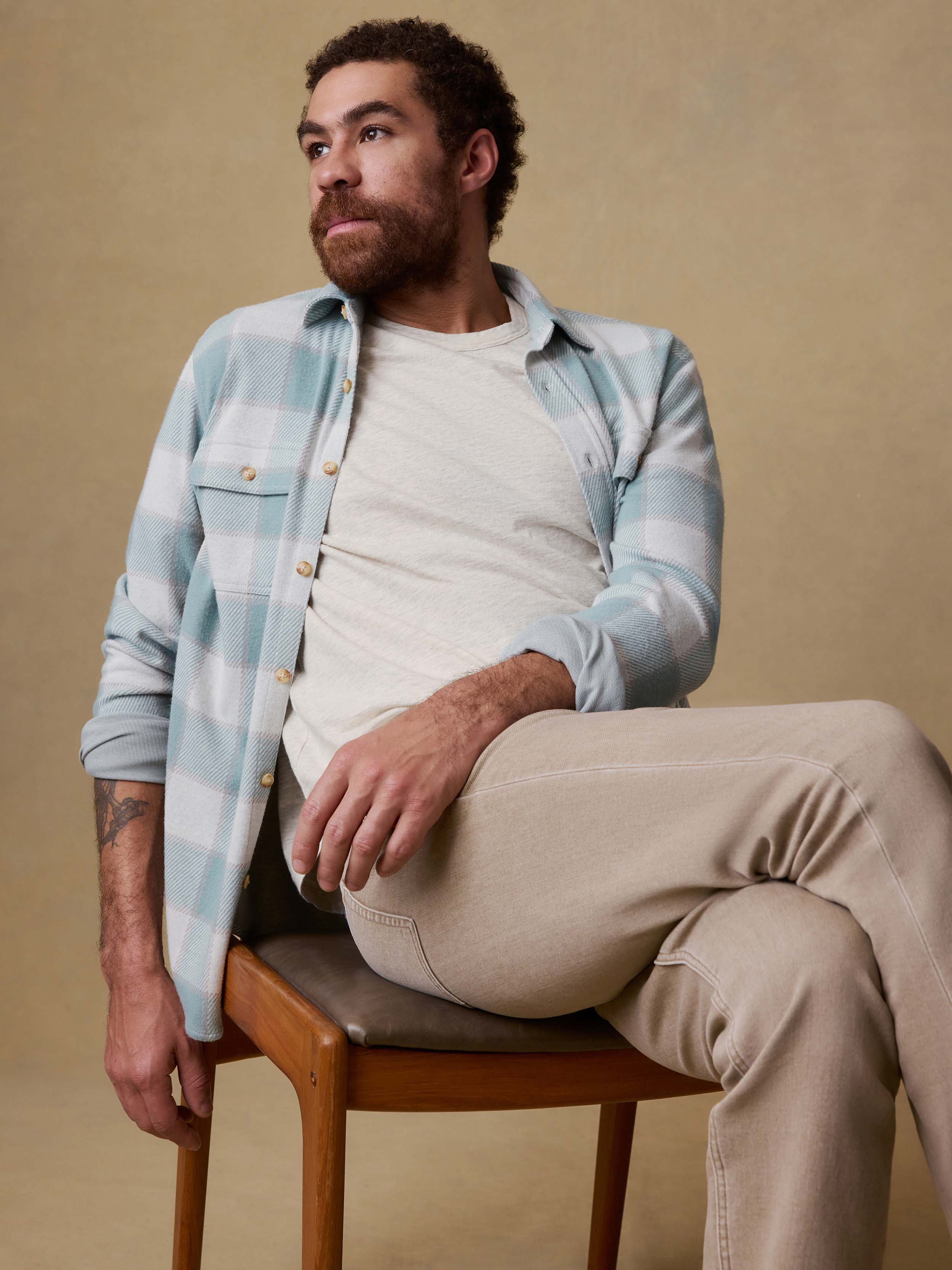 Man sitting on a chair wearing a light blue plaid shirt over a white t-shirt and beige pants.
