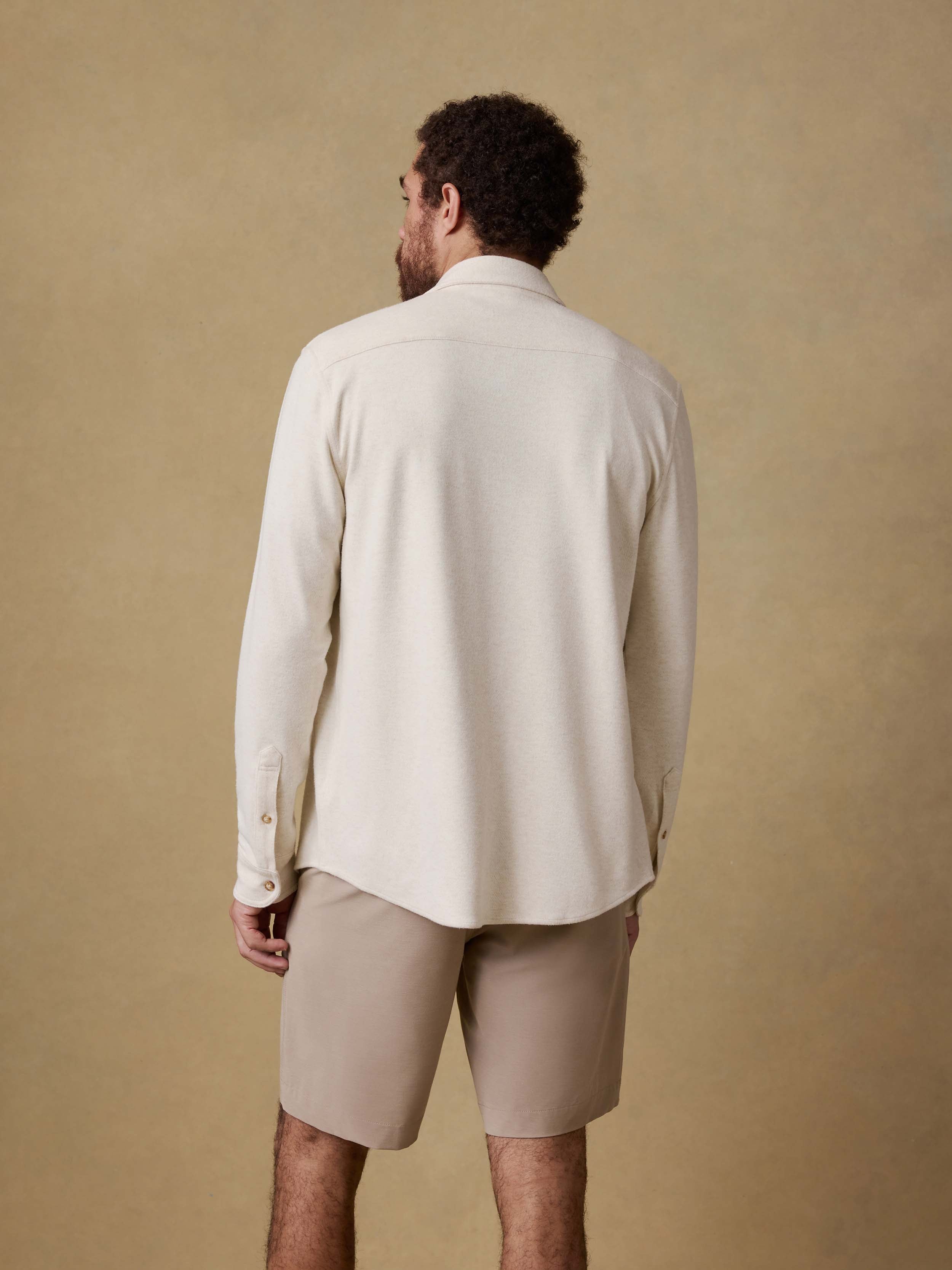Back view of a man wearing an off white button up shirt and beige shorts against a beige background