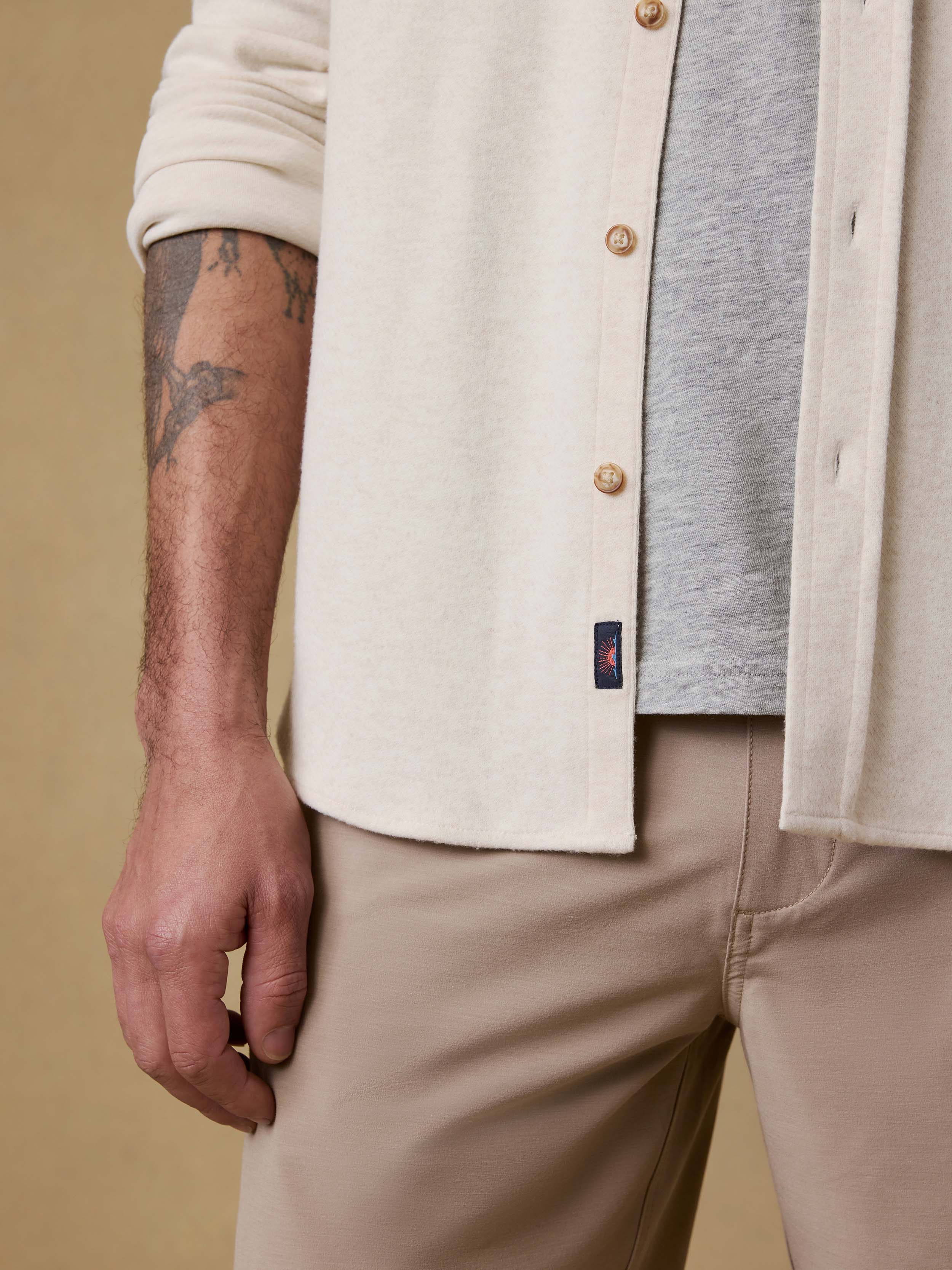 Detail shot of a man wearing an off white button up shirt with a visible Faherty brand logo over a gray t-shirt and beige shorts against beige background
