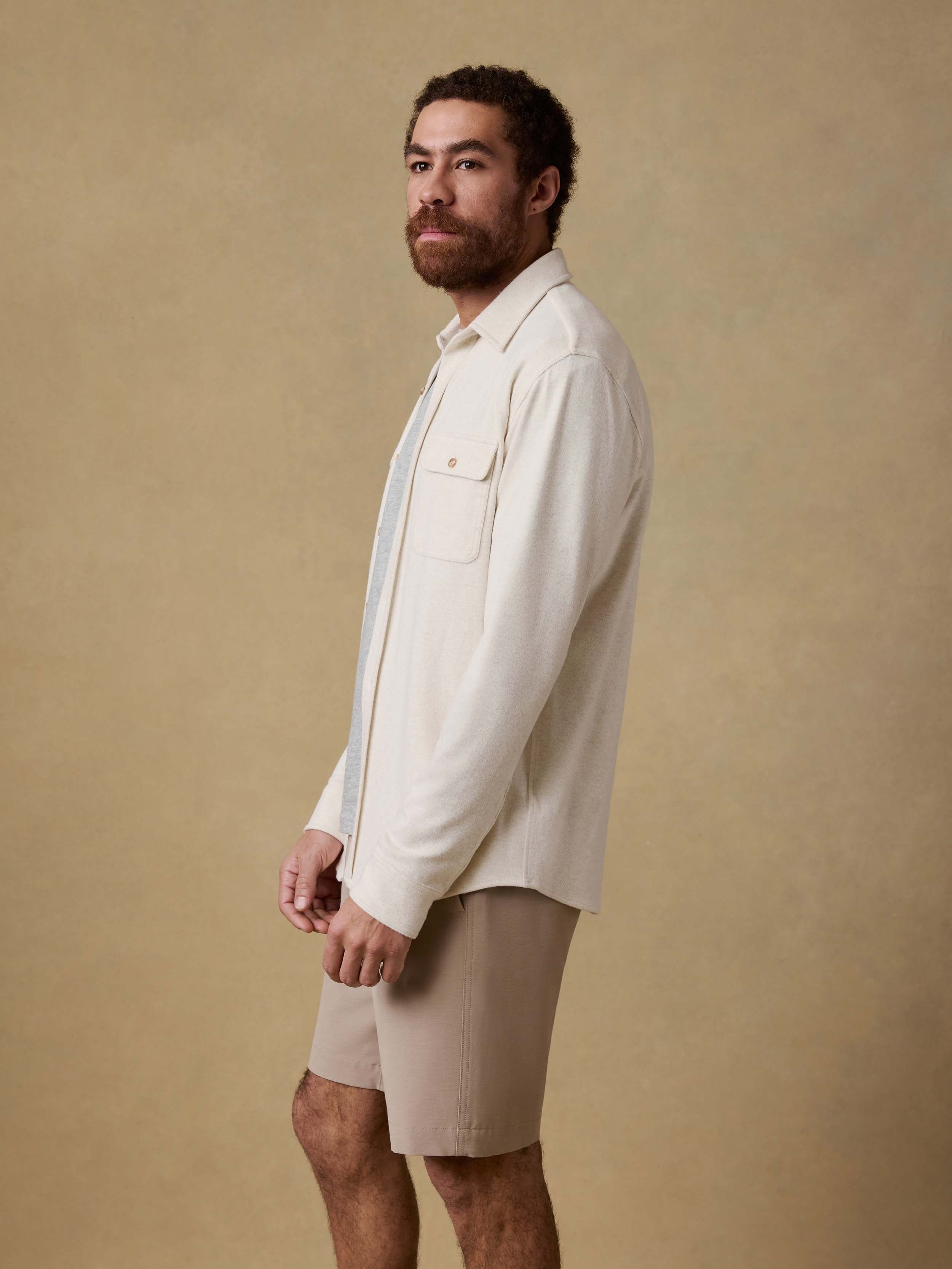 Man wearing an off white button up shirt over a gray t-shirt and beige shorts against a beige background