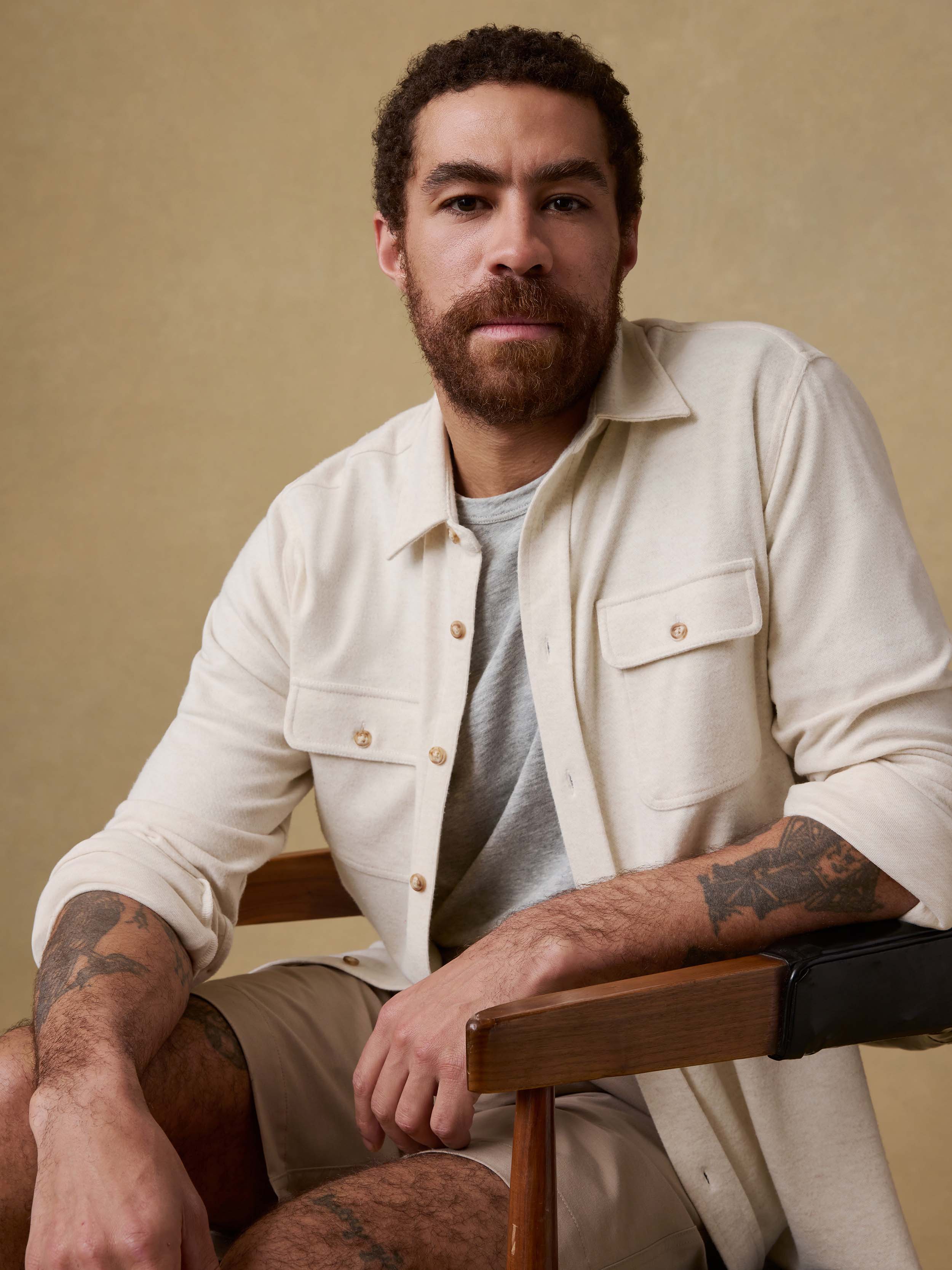 Man sitting on a chair wearing an off white button up shirt over a gray t-shirt and beige shorts against a beige background