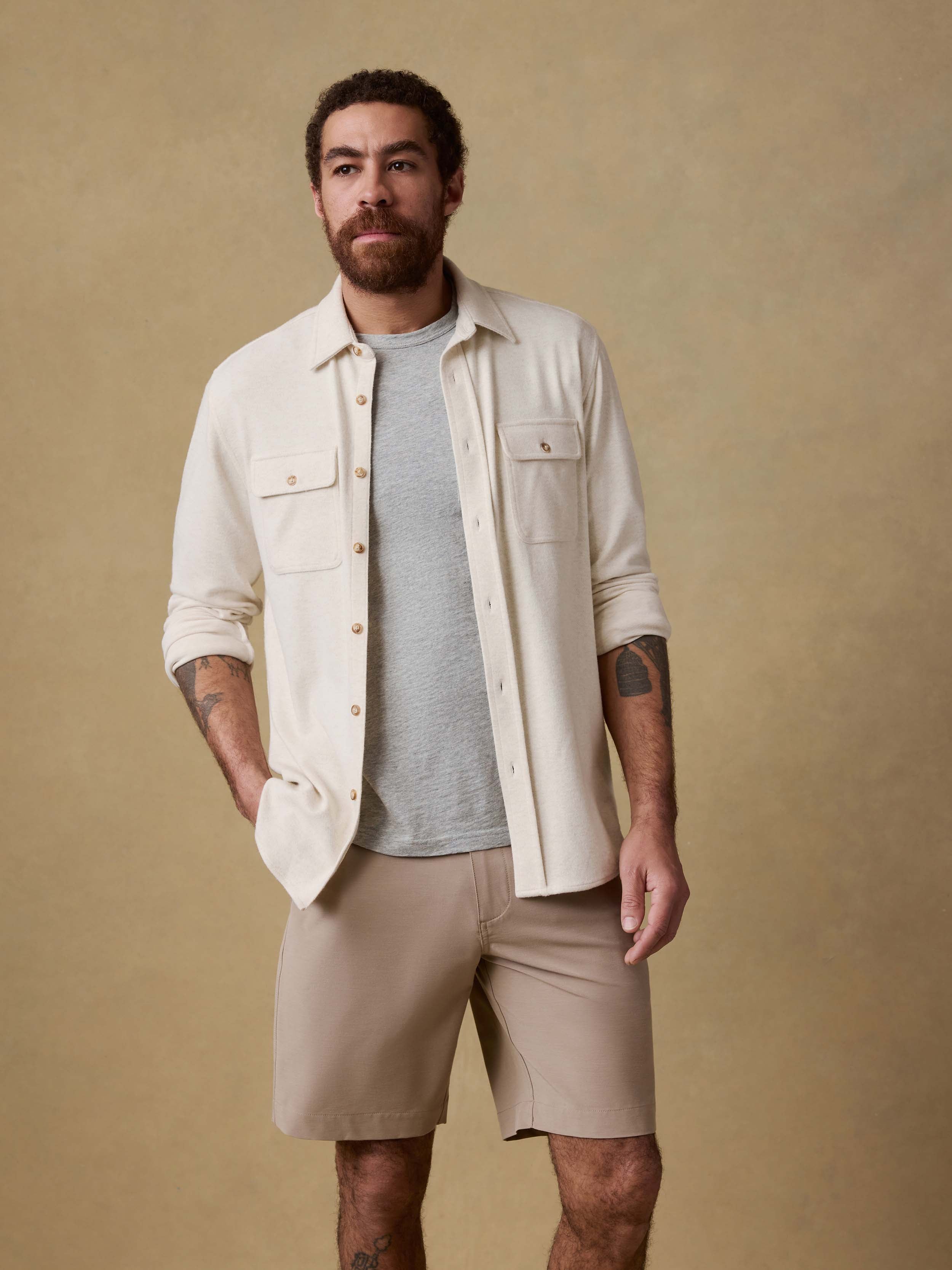 Man wearing an off white button up shirt over a gray t-shirt and beige shorts against a beige background