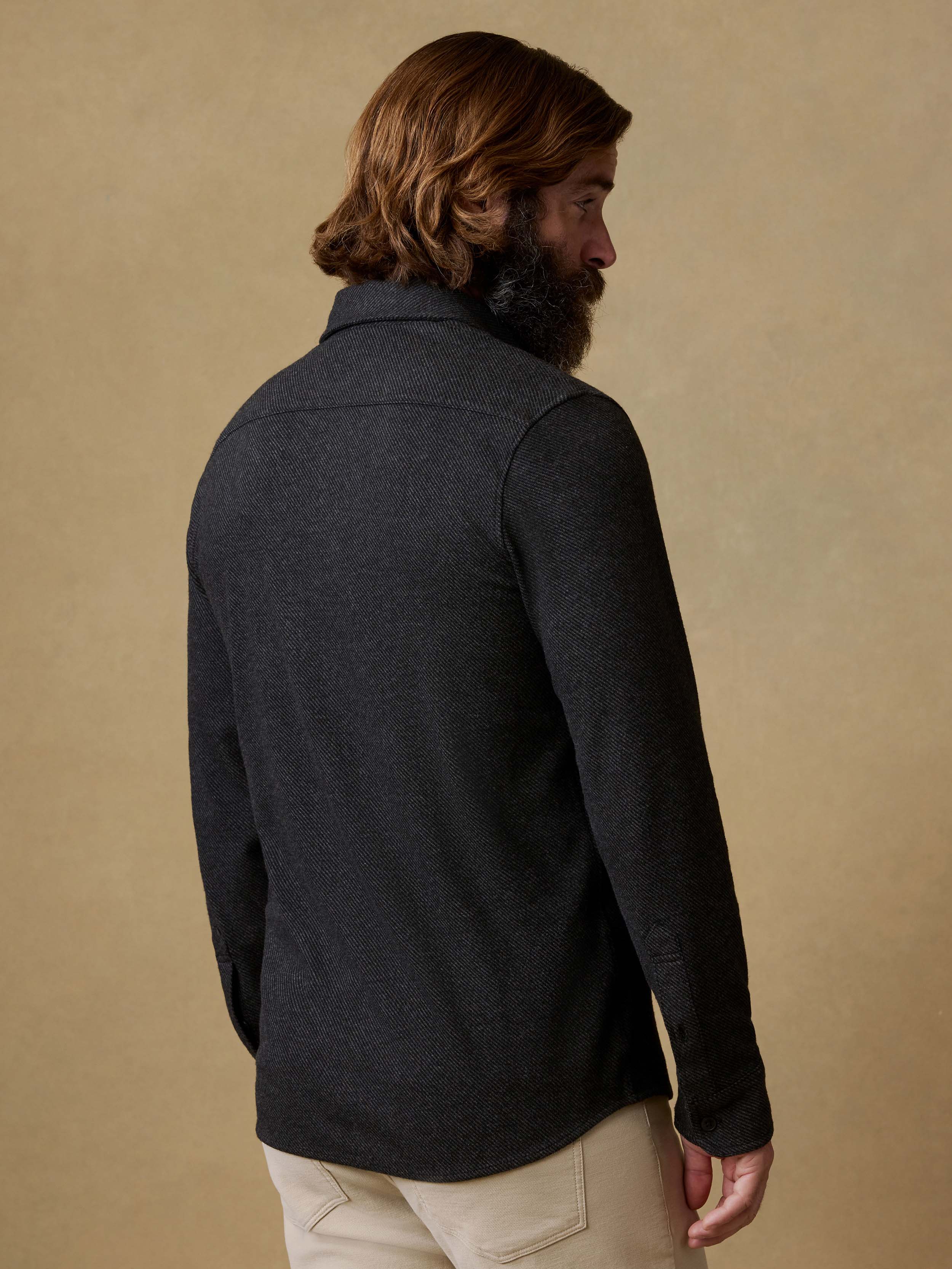 Back view of a man wearing a black button-up shirt and beige pants against a beige background