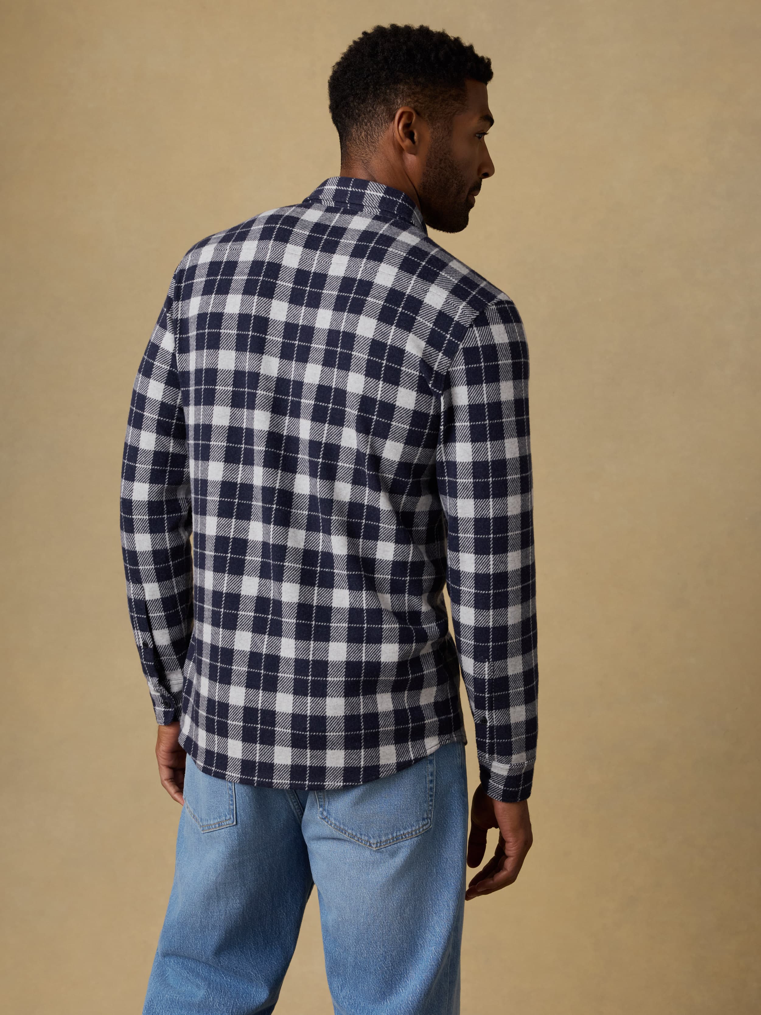 Back view of a man wearing a navy and white checkered long sleeve button up shirt and blue jeans against a beige background