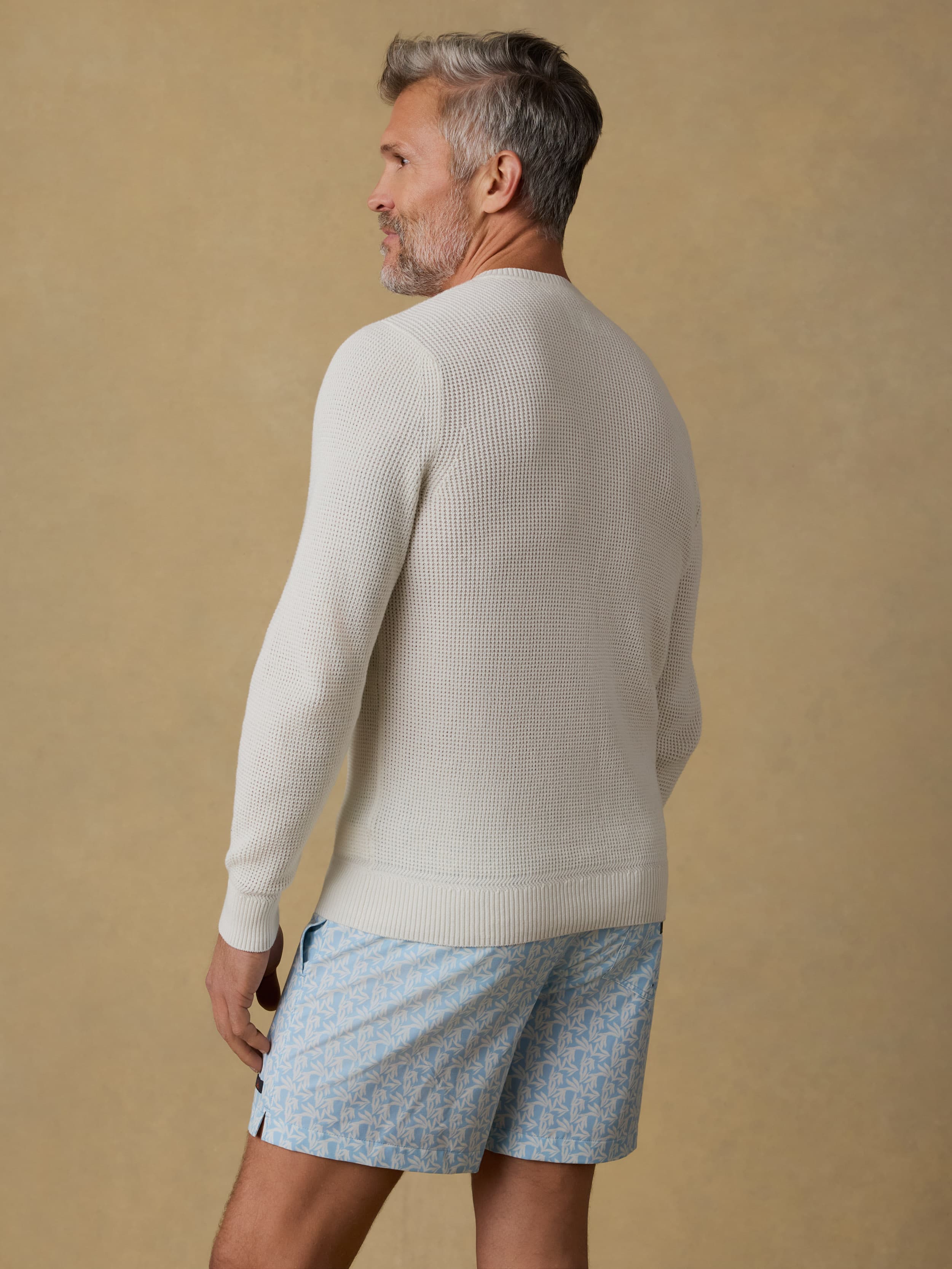 Back view of a man wearing a white crewneck sweater and patterned swim trunks against a beige background