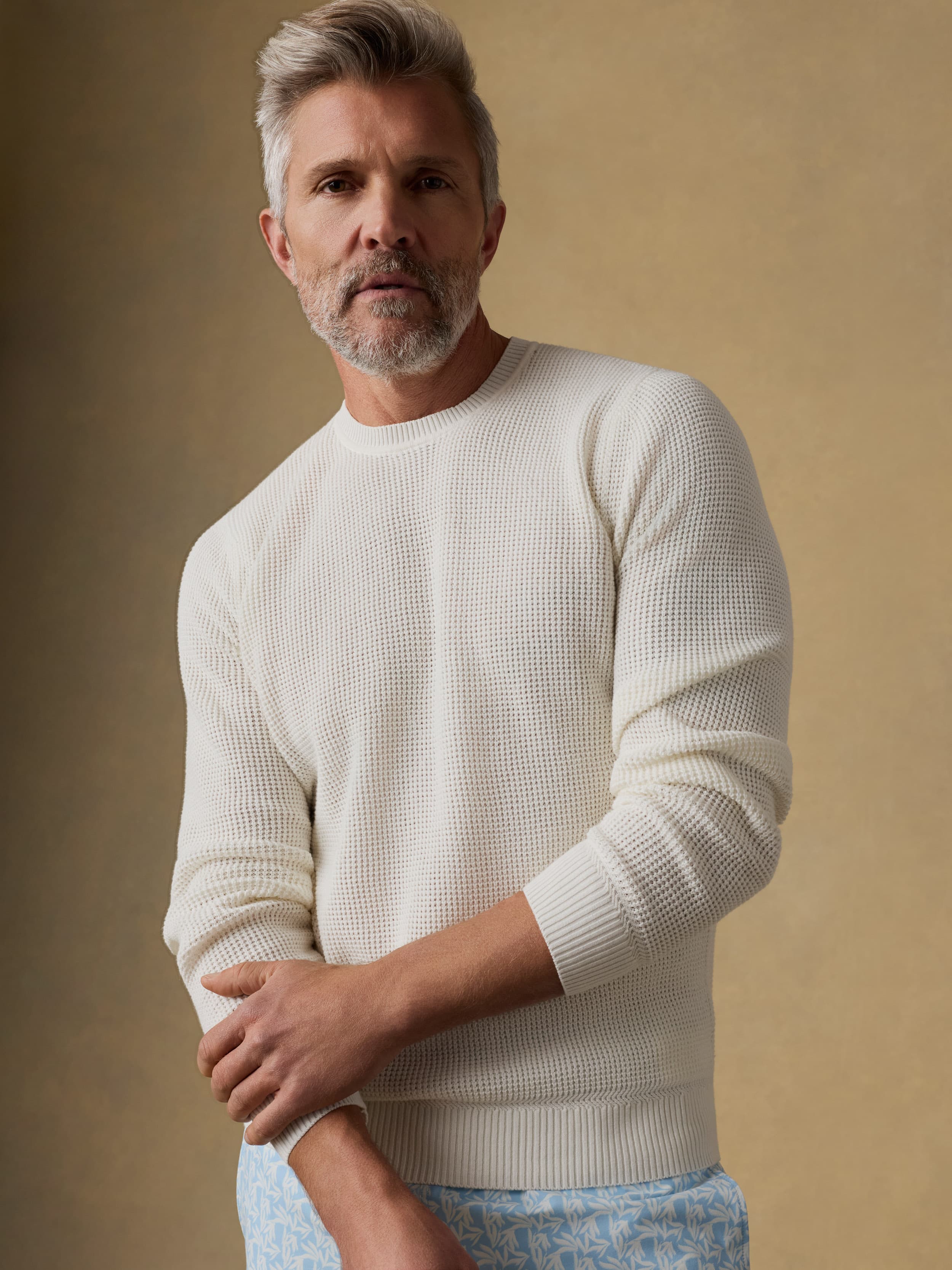 Man wearing a white crewneck sweater and patterned swim trunks against a beige background