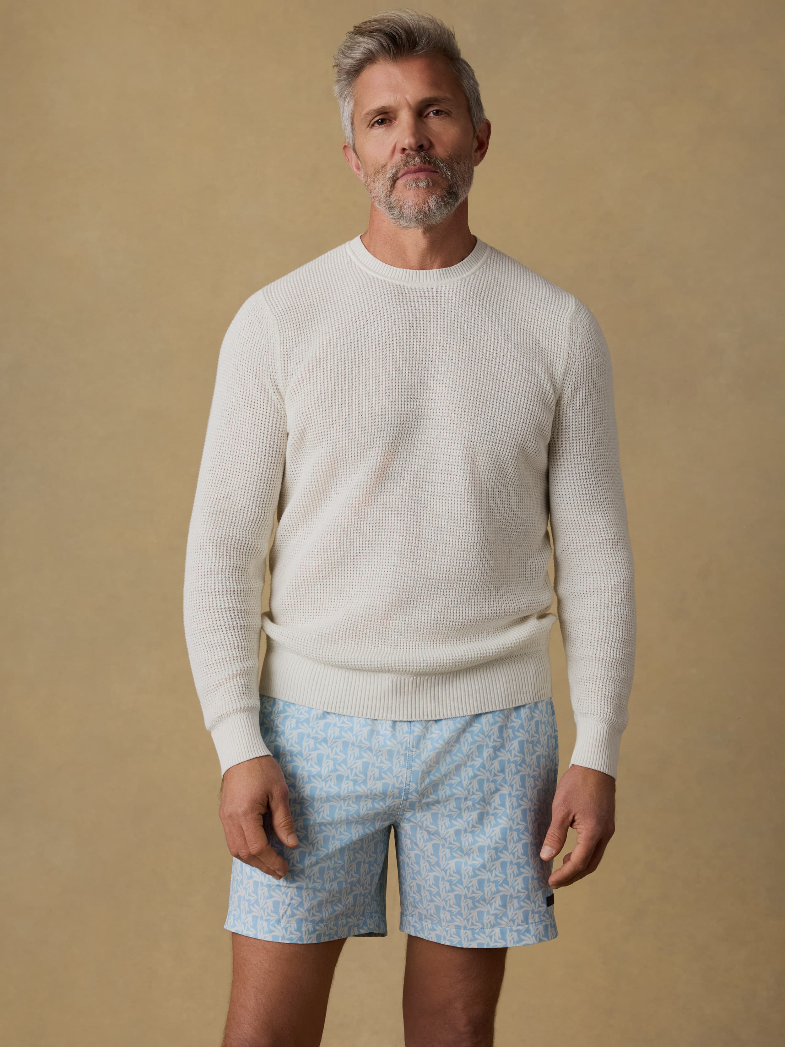 Man wearing a white crewneck sweater and patterned swim trunks against a beige background