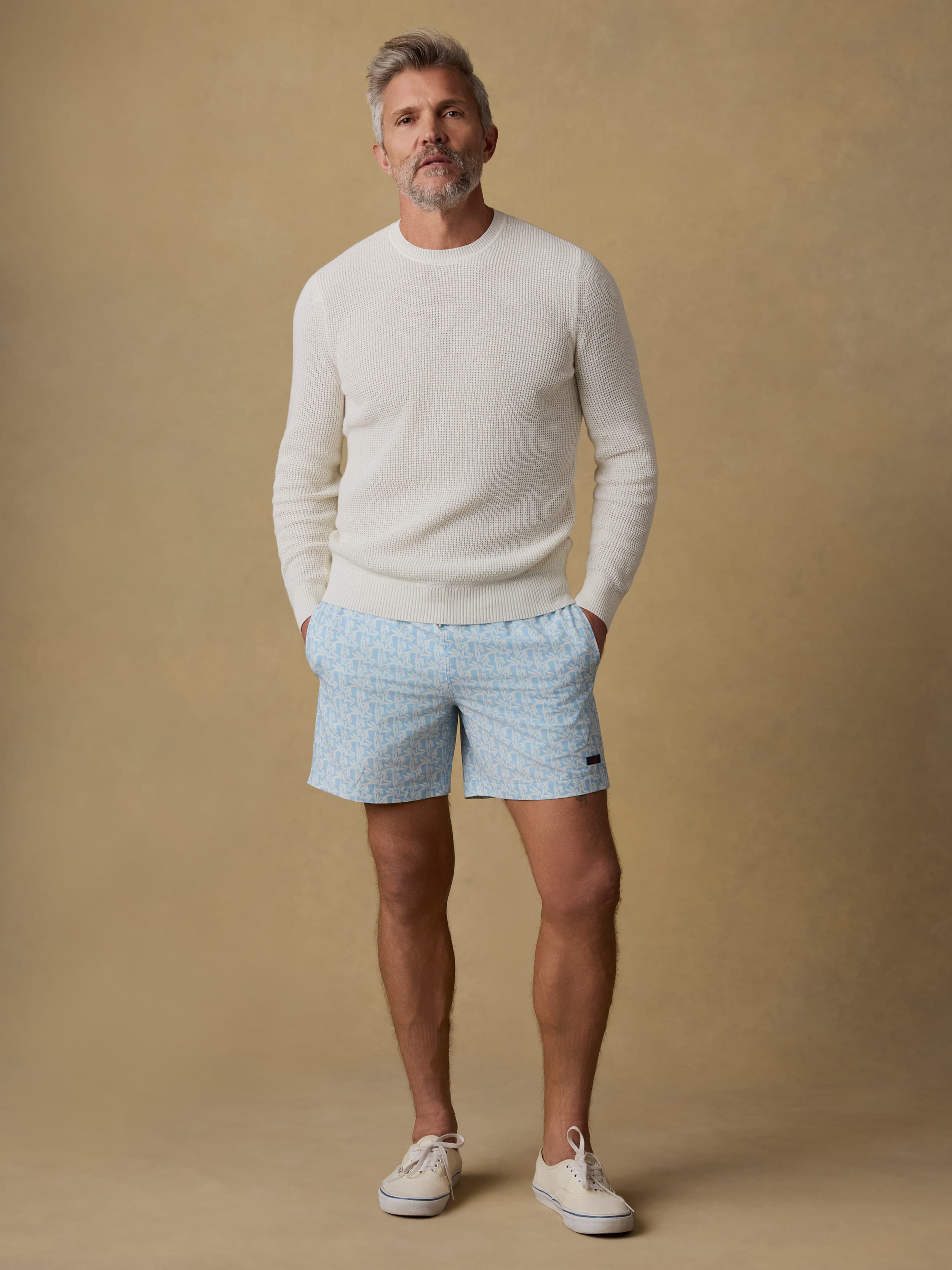 Man wearing a white crewneck sweater and patterned swim trunks against a beige background