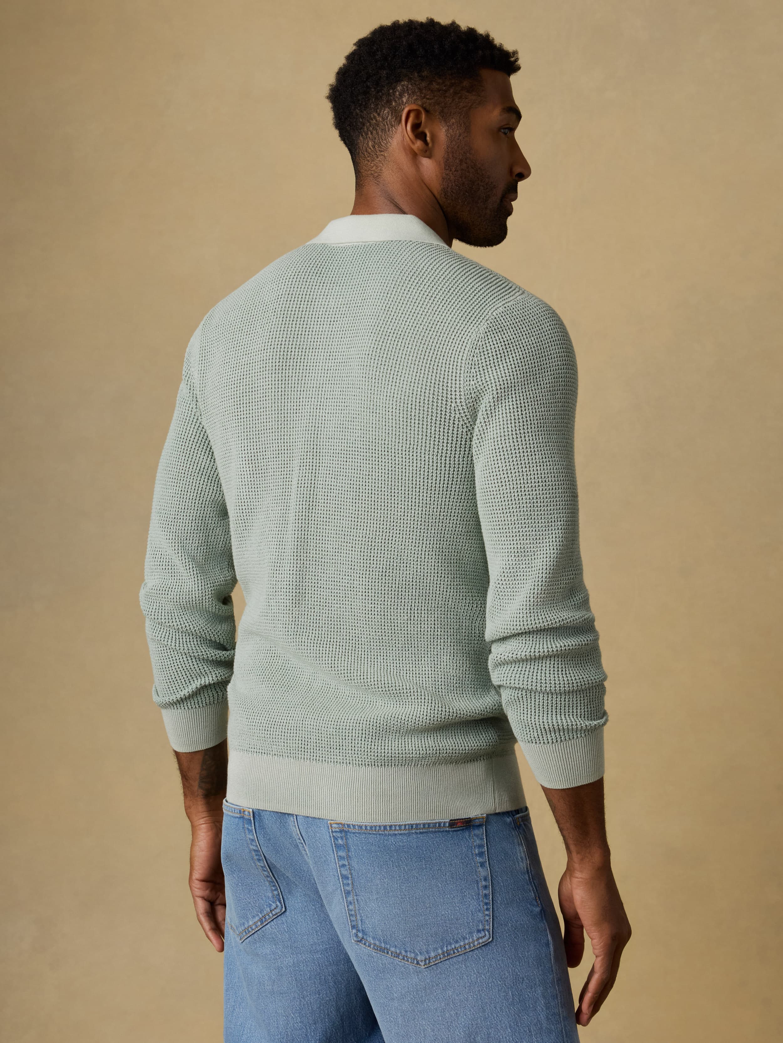 Back view of a man wearing a light green polo sweater with blue jeans on a beige background