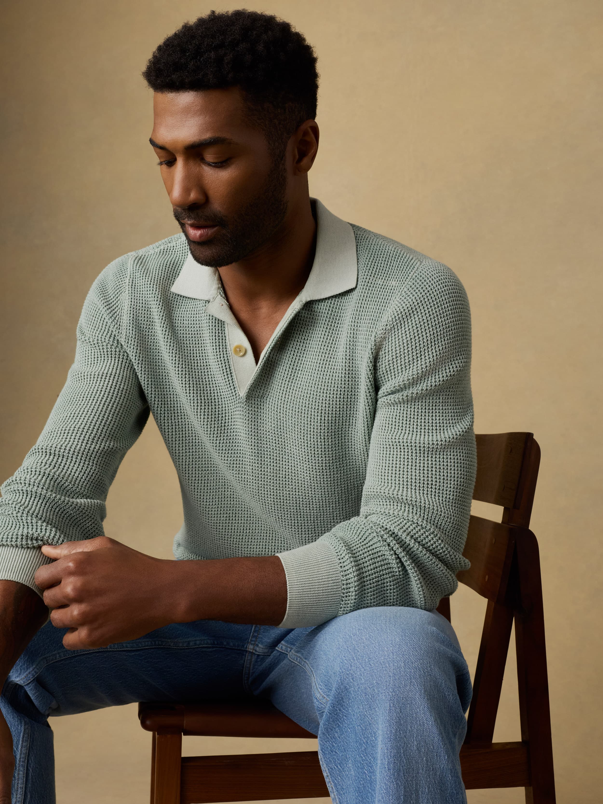 Man sitting on a wooden chair wearing a light green polo sweater with blue jeans on a beige background