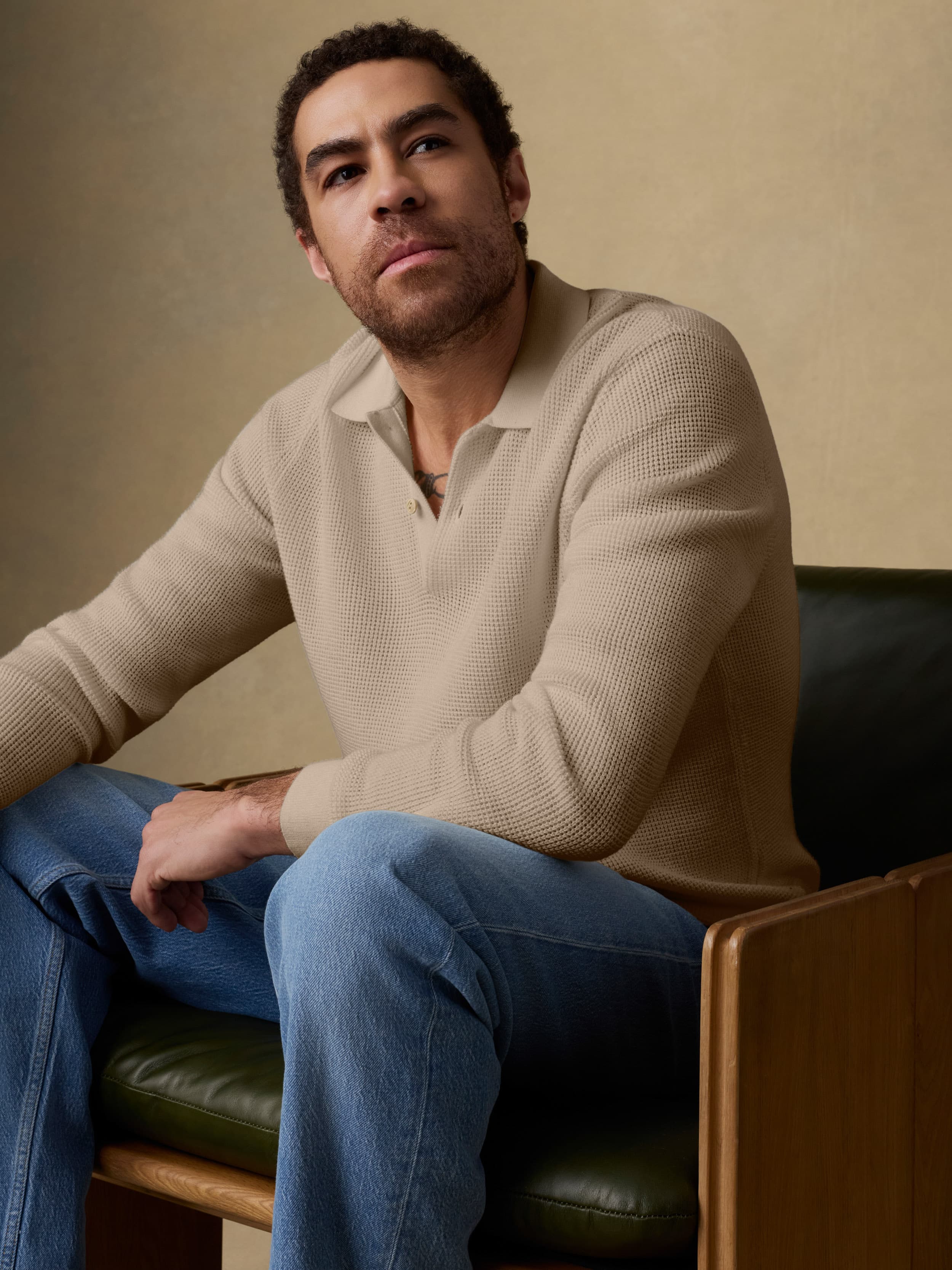 Man sitting on a wooden chair wearing a beige sweater and blue jeans against a beige background
