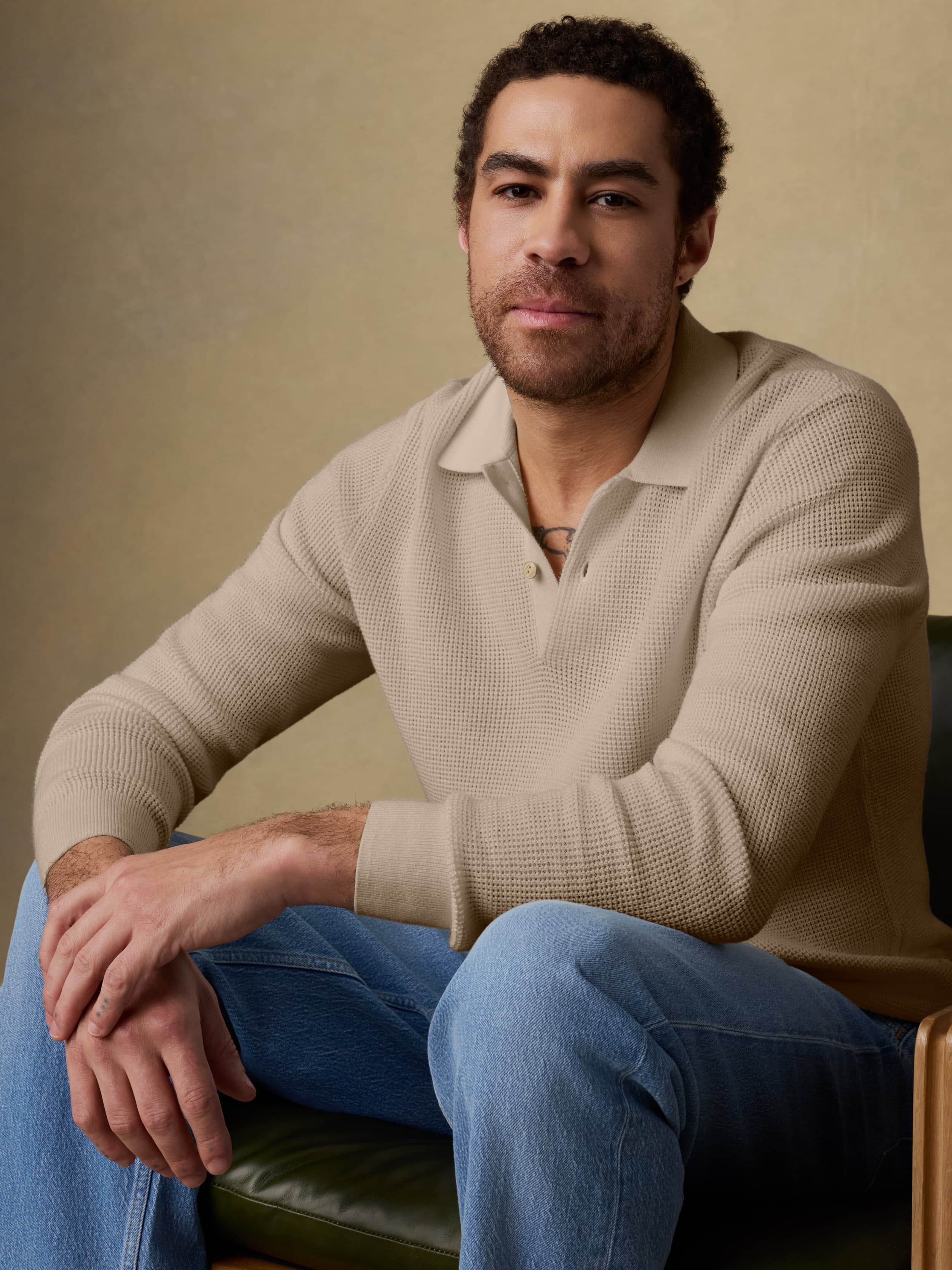 Man sitting on a wooden chair wearing a beige sweater and blue jeans against a beige background