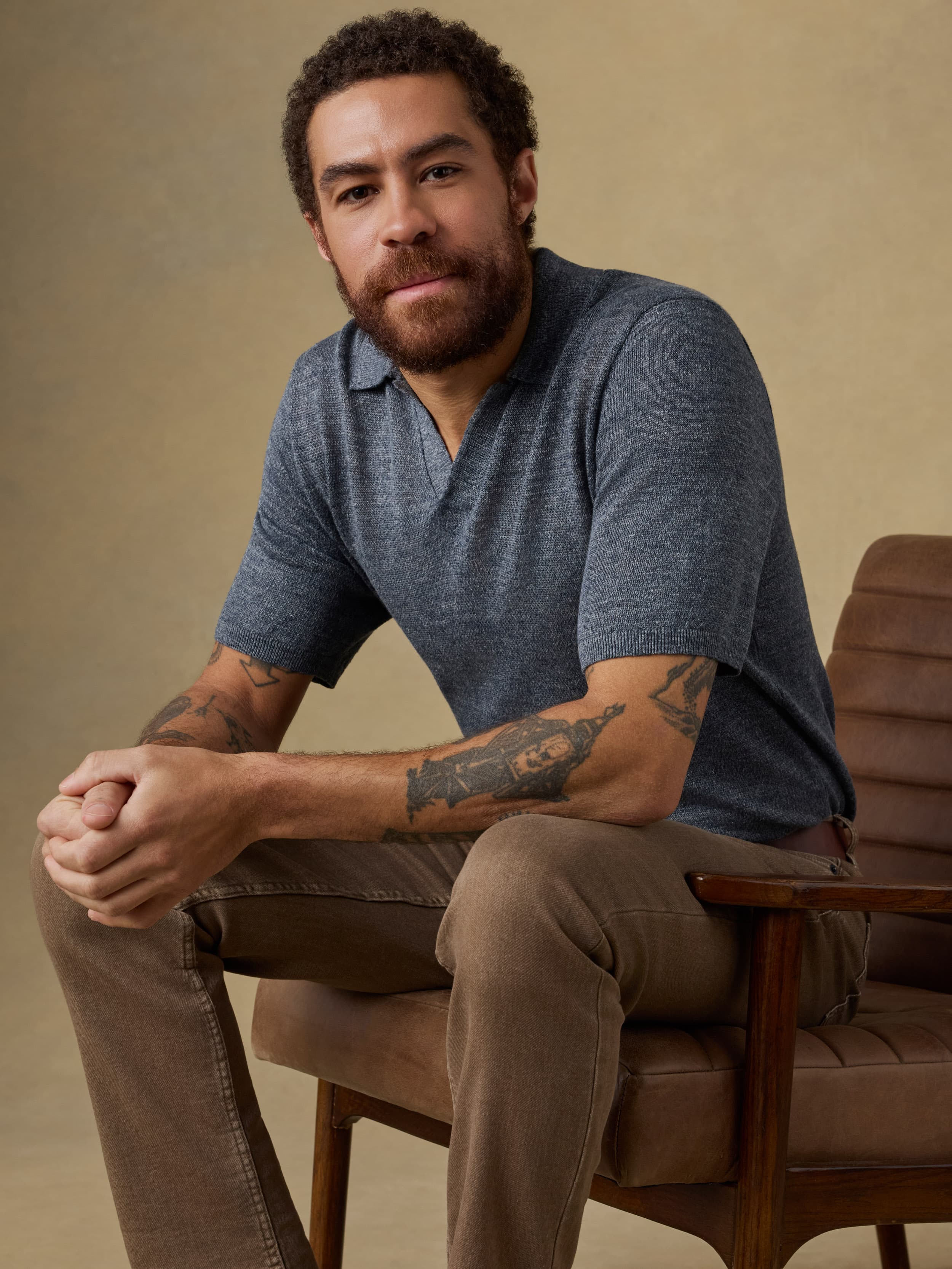 Man sitting on a wooden chair wearing a blue polo shirt and brown pants against a beige background