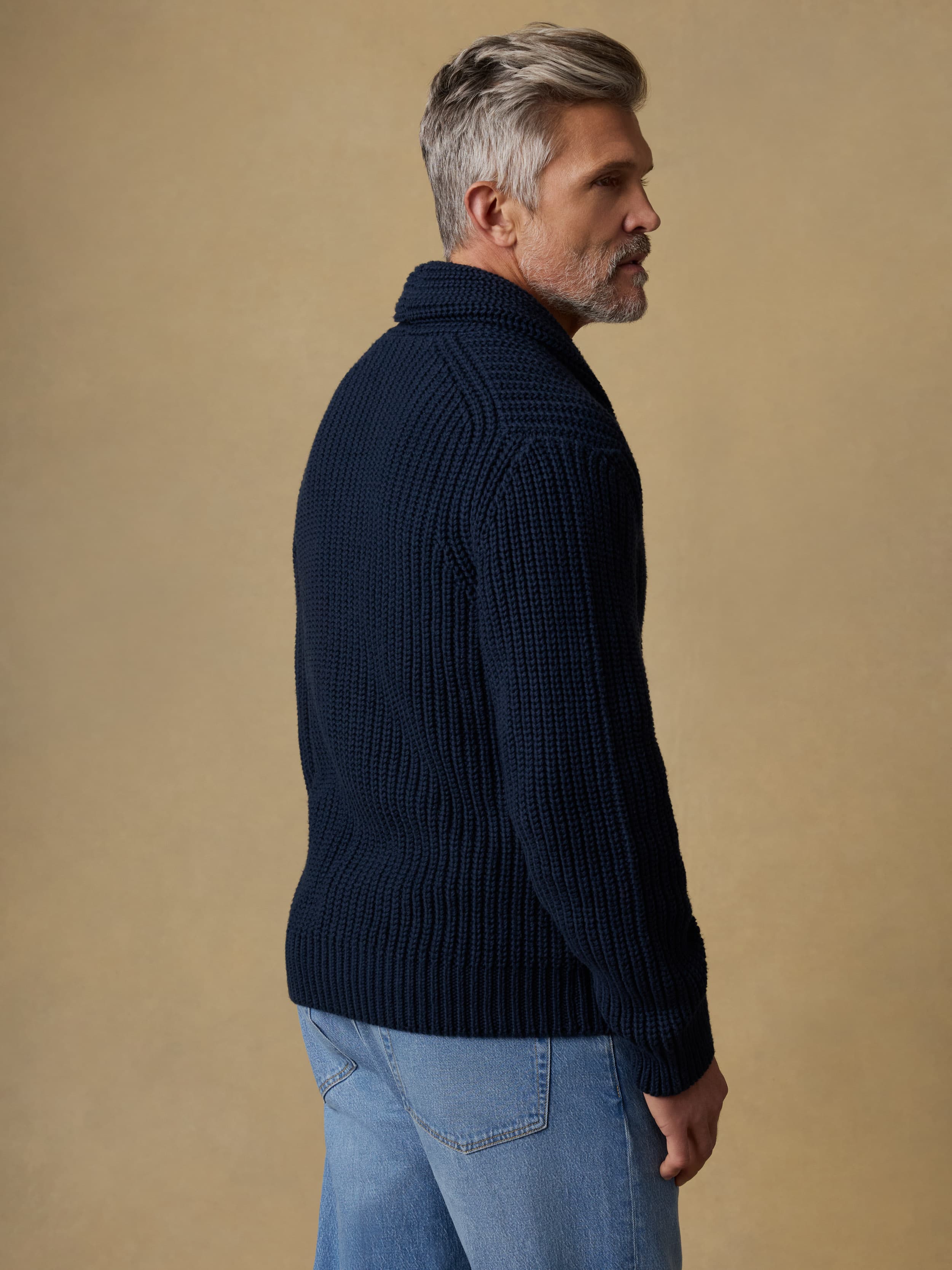Back view of a man wearing a navy blue cardigan and blue jeans against a beige background