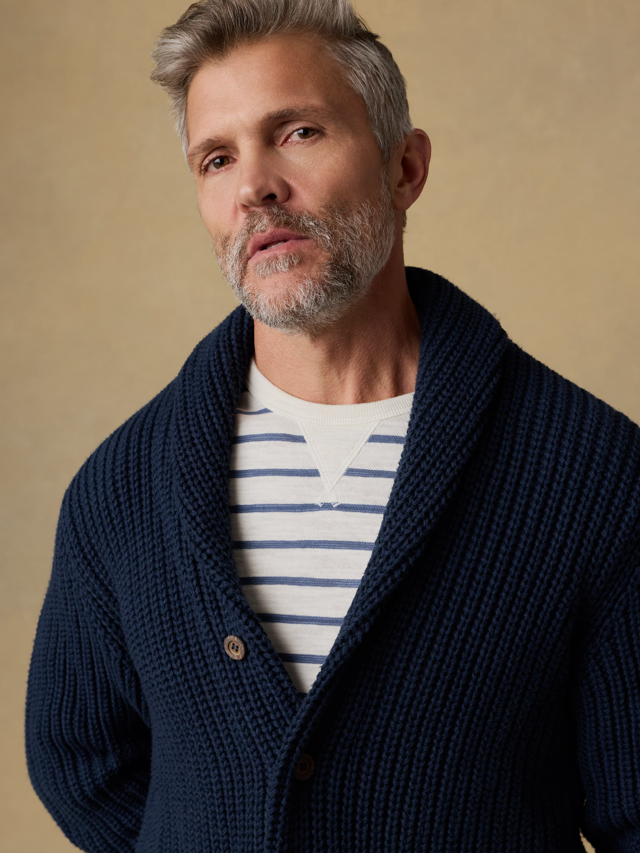 Man wearing a navy blue cardigan over a blue and white striped crewneck against a beige background