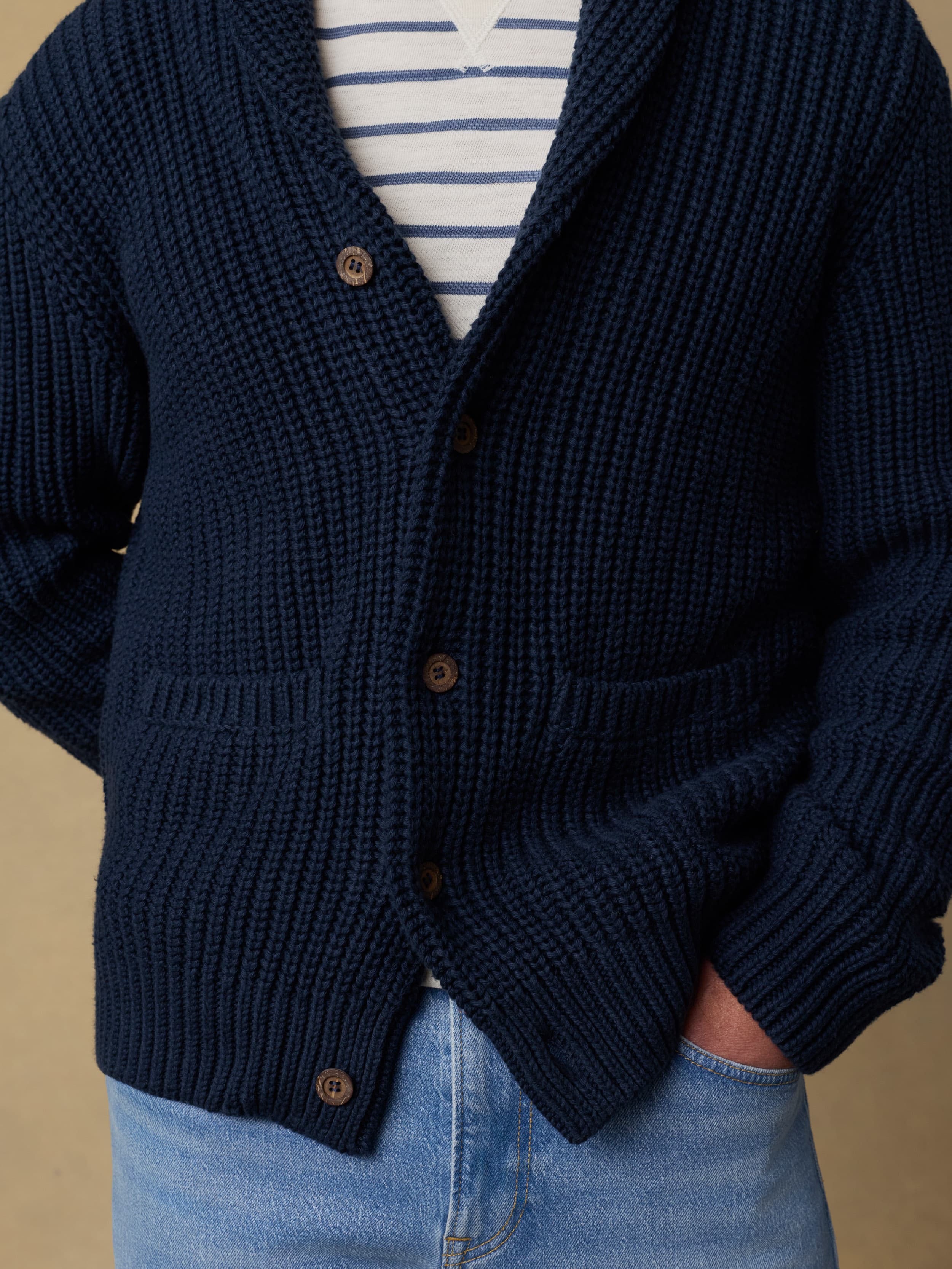 Detail shot of a man wearing a navy blue cardigan over a blue and white striped crewneck and blue jeans against a beige background