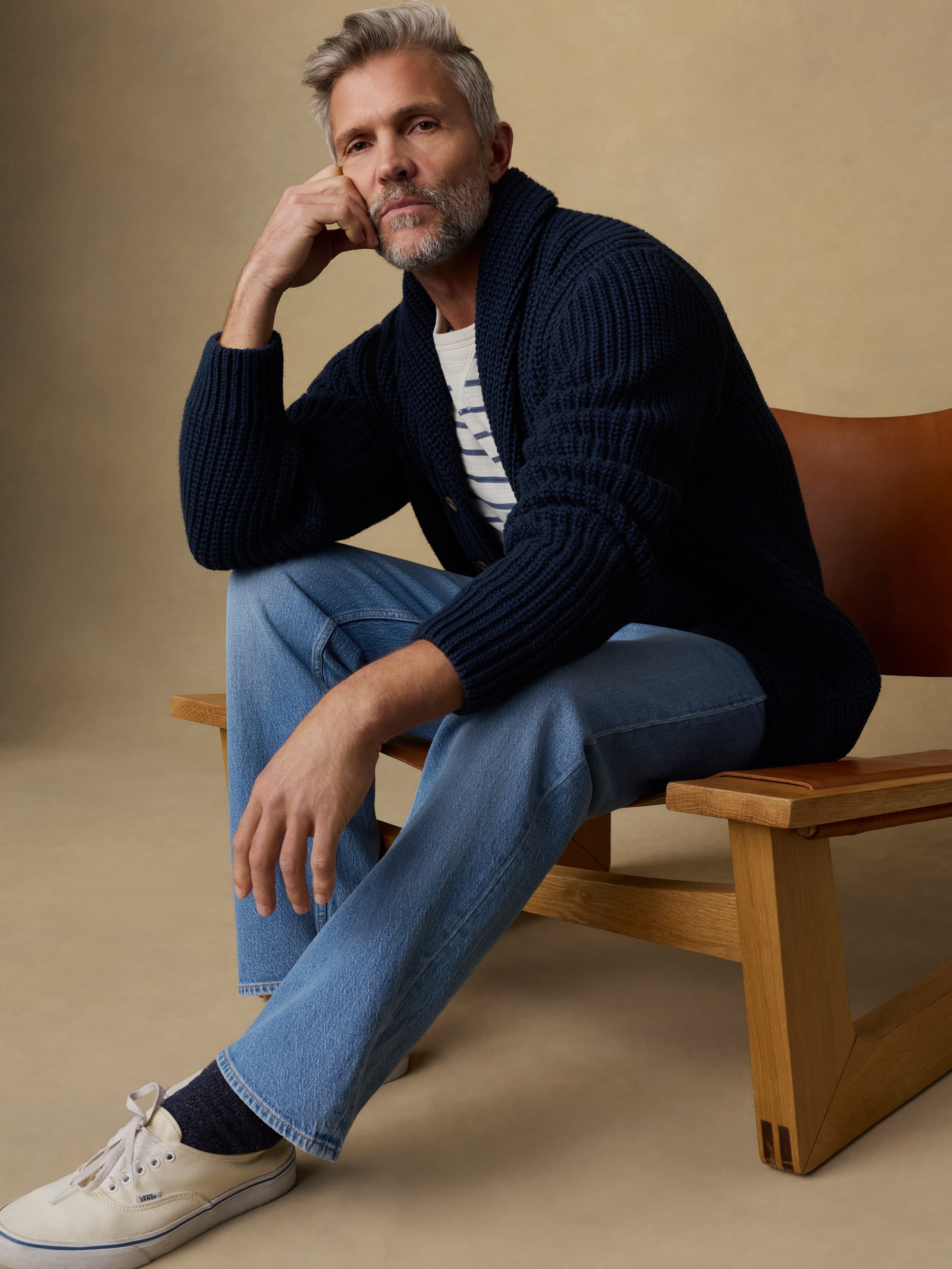 Man sitting on a wooden chair wearing a navy blue cardigan over a blue and white striped crewneck and blue jeans against a beige background