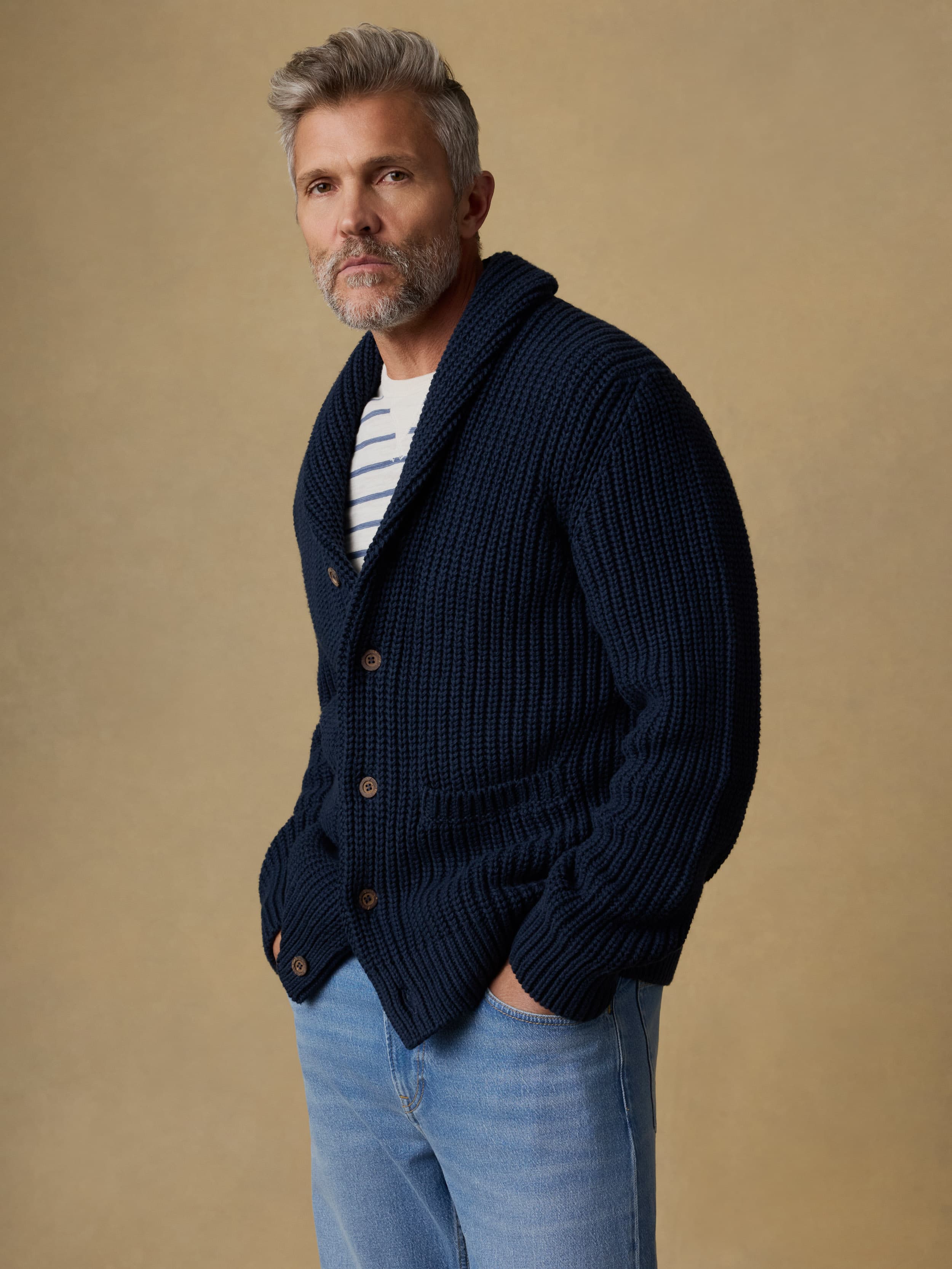 Man wearing a navy blue cardigan over a blue and white striped crewneck and blue jeans against a beige background