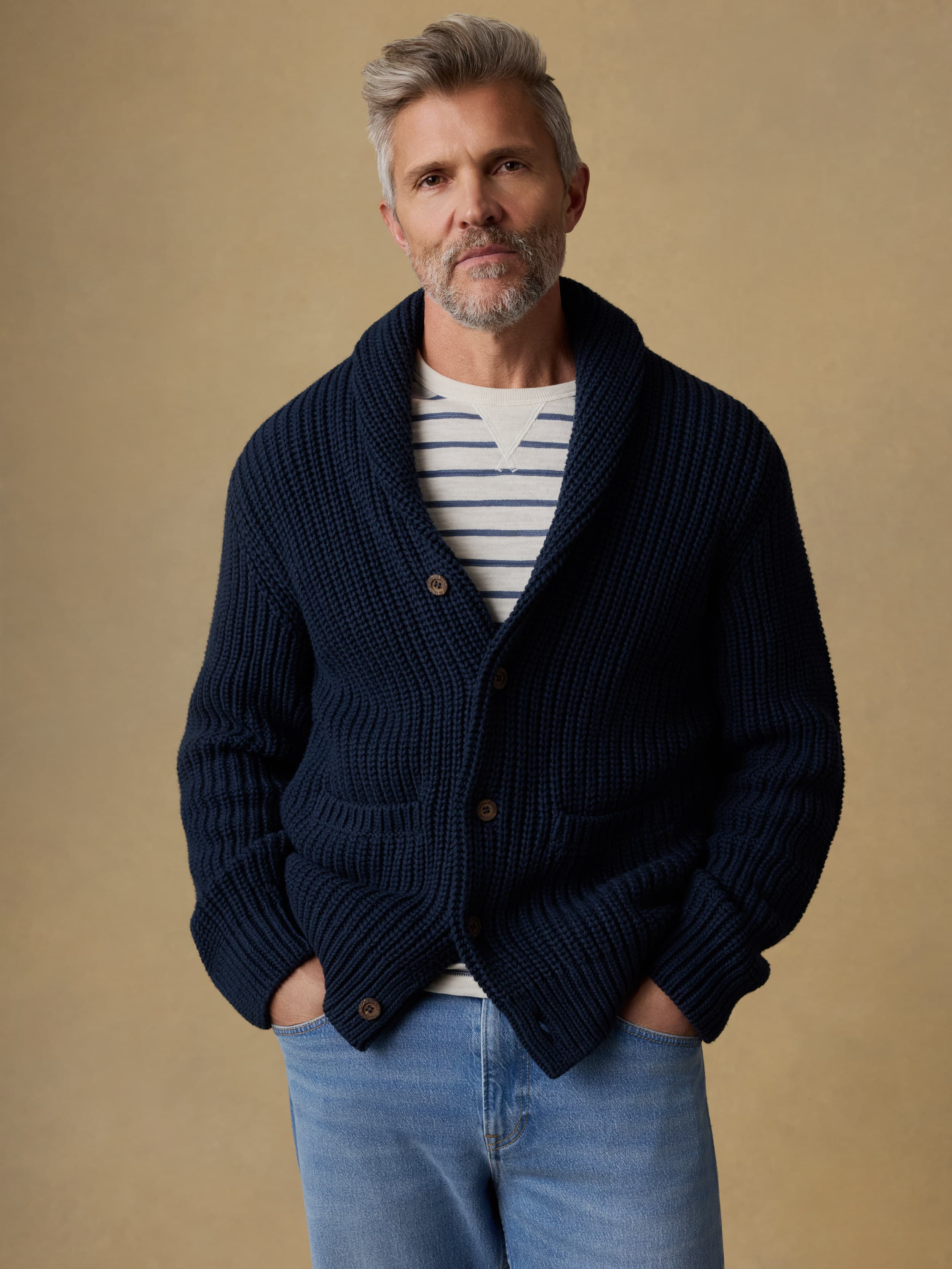 Man wearing a navy blue cardigan over a blue and white striped crewneck and blue jeans against a beige background