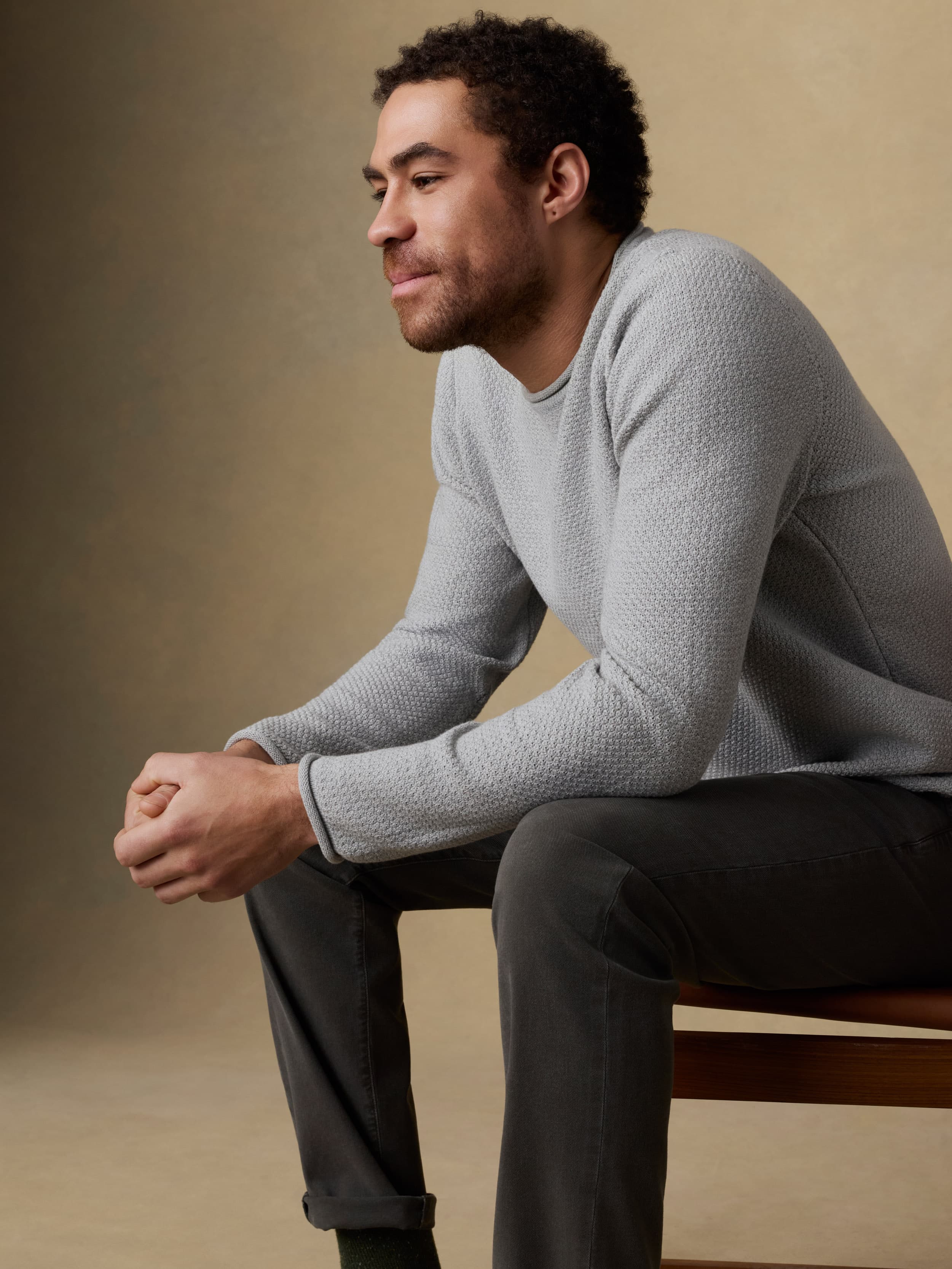 Man sitting on a wooden chair wearing a gray sweater and black jeans against a beige background