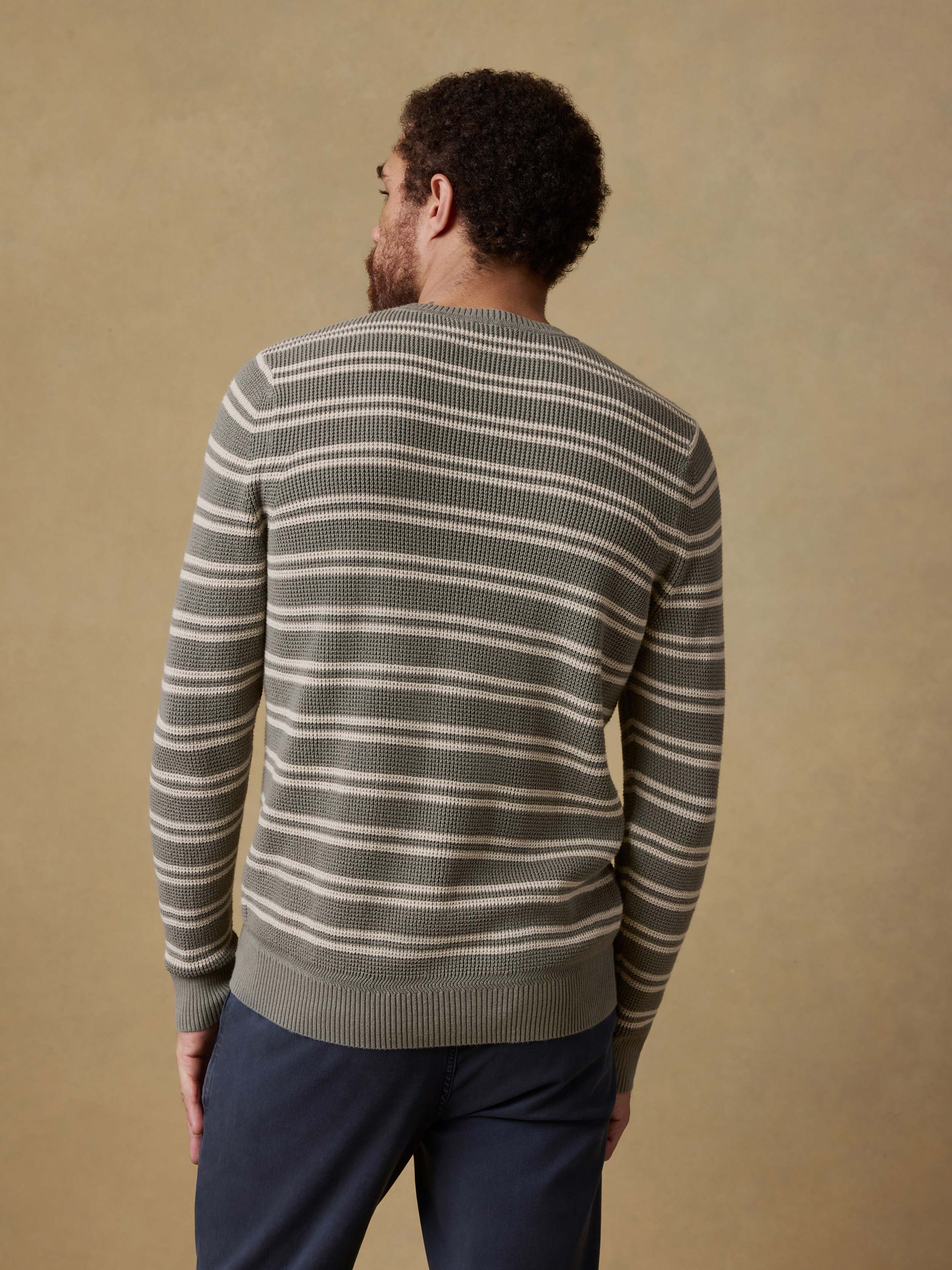 Back view of a man wearing a green striped sweater and navy pants against a beige background